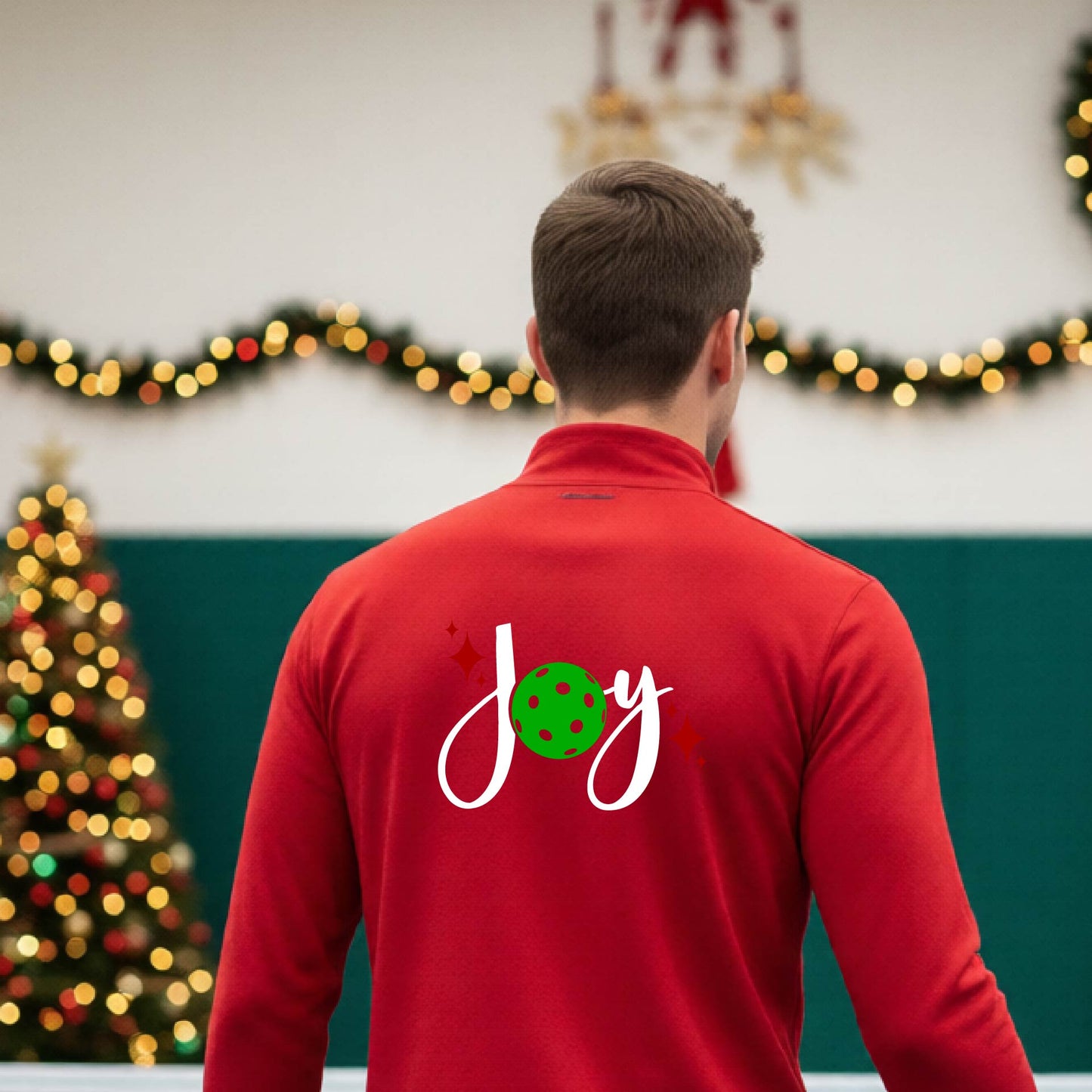 A man viewed from behind wearing a red quarter-zip pullover with a festive Christmas graphic on the back that reads 'Joy' in white script with a green pickleball and red stars. The photo is taken inside a gymnasium decorated for Christmas.