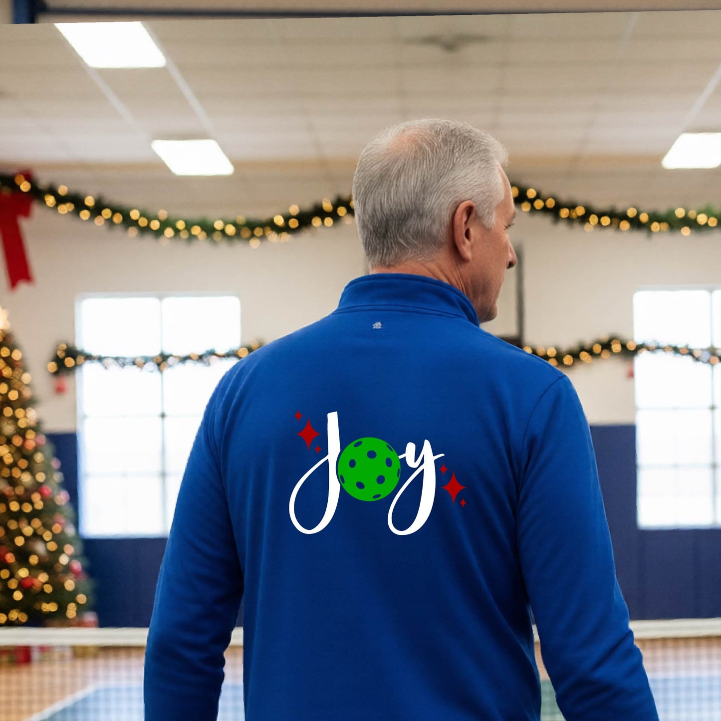 A man viewed from behind wearing a royal blue quarter-zip pullover with a festive Christmas graphic on the back that reads 'Joy' in white script with a green pickleball and red stars. The photo is taken inside a gymnasium decorated for Christmas.