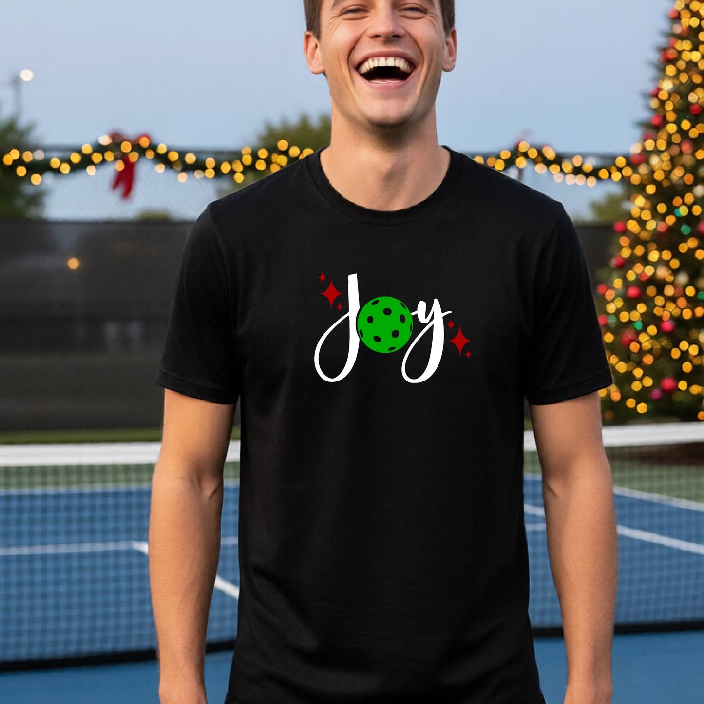 A man laughing on a pickleball court wearing a black short-sleeve shirt with a 'JOY' holiday graphic with the O replaced by a green pickleball wearing a Santa hat. A decorated Christmas tree and warm string lights are blurred in the background