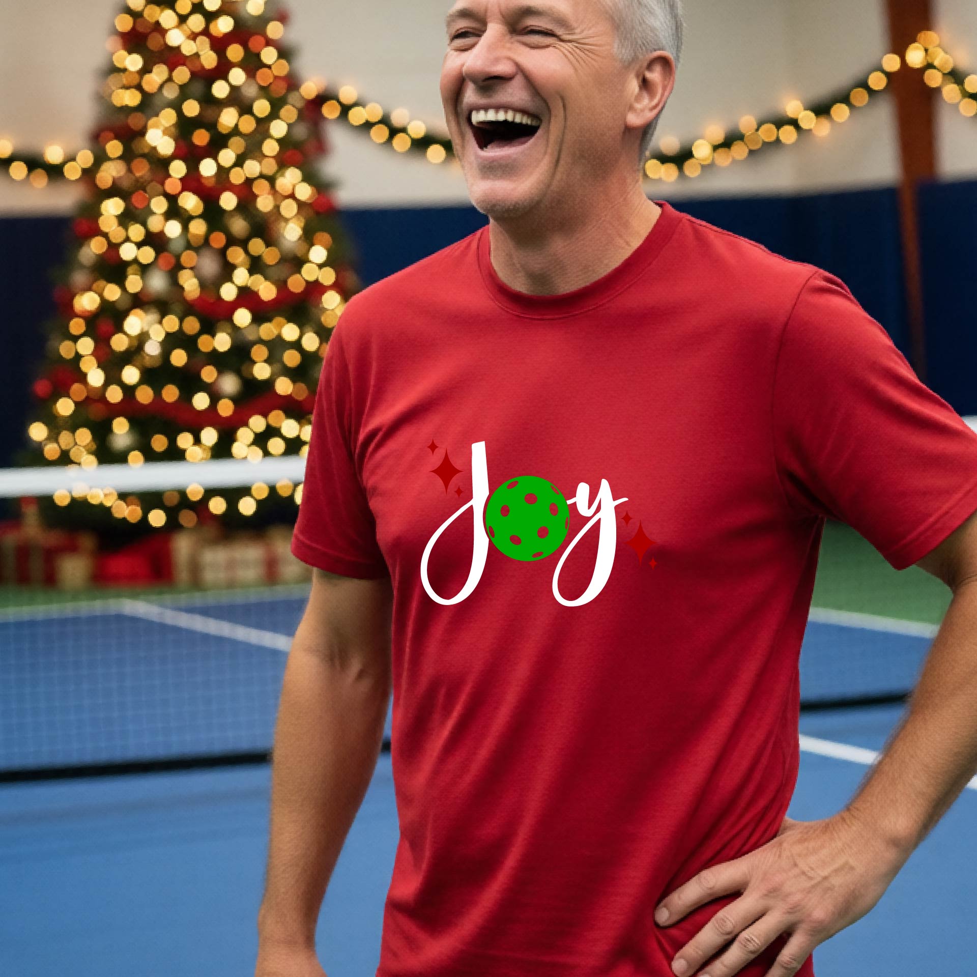 A man laughing on a pickleball court wearing a red short-sleeve shirt with a 'JOY' holiday graphic with the O replaced by a green pickleball wearing a Santa hat. A decorated Christmas tree and warm string lights are blurred in the background