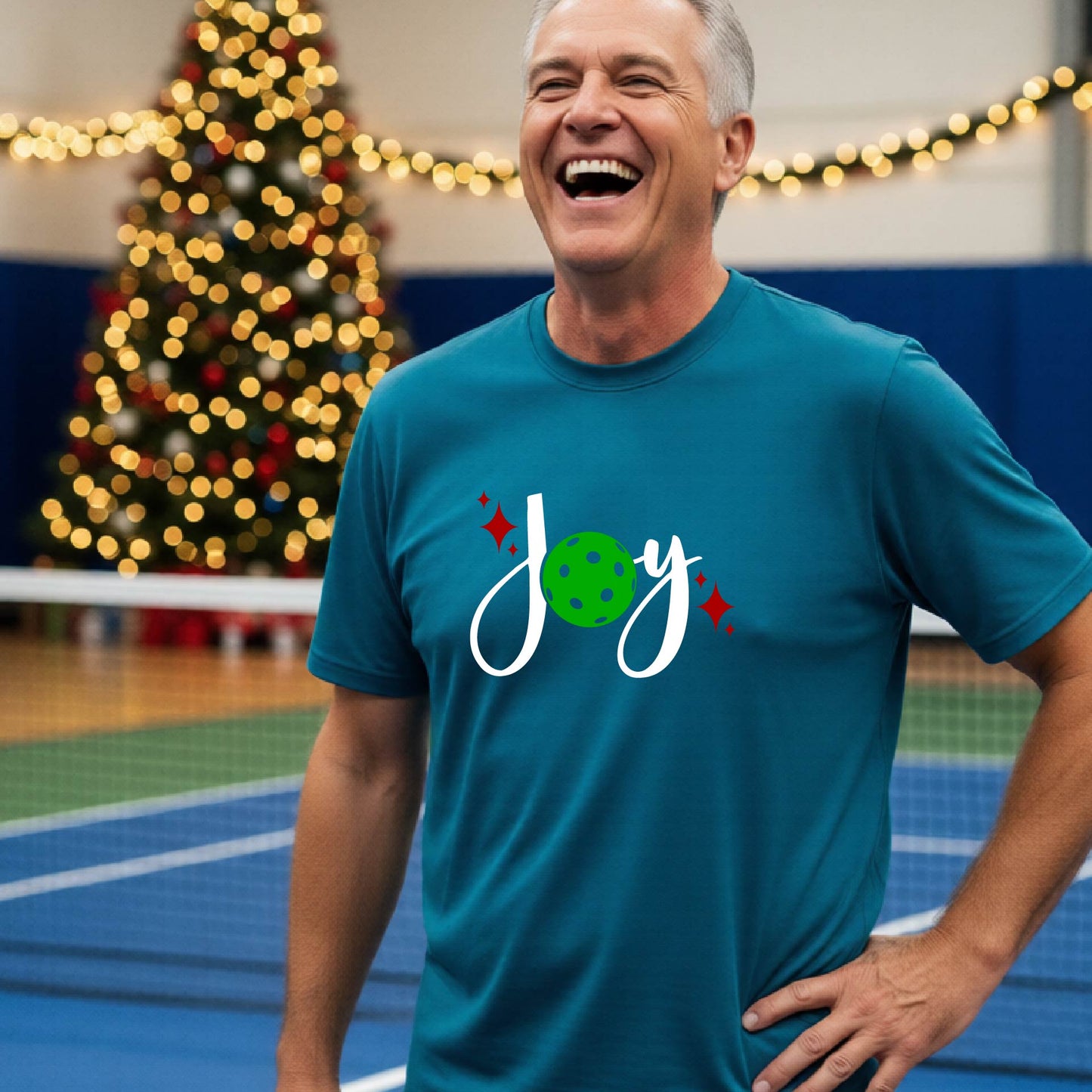 A man laughing on a pickleball court wearing a teal short-sleeve shirt with a 'JOY' holiday graphic with the O replaced by a green pickleball wearing a Santa hat. A decorated Christmas tree and warm string lights are blurred in the background
