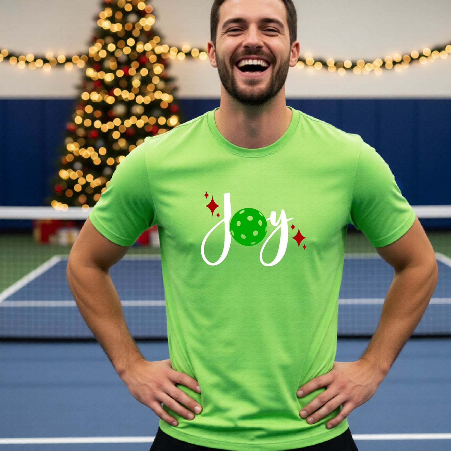 A man laughing on a pickleball court wearing a lime green short-sleeve shirt with a 'JOY' holiday graphic with the O replaced by a green pickleball wearing a Santa hat. A decorated Christmas tree and warm string lights are blurred in the background
