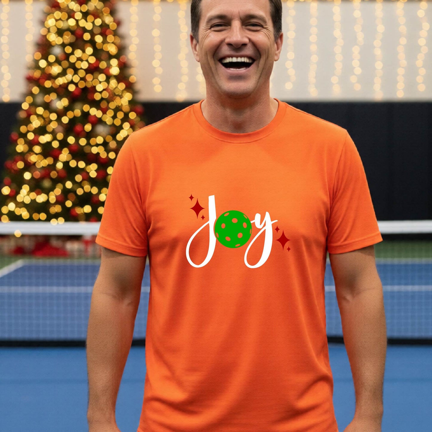 A man laughing on a pickleball court wearing an orange short-sleeve shirt with a 'JOY' holiday graphic with the O replaced by a green pickleball wearing a Santa hat. A decorated Christmas tree and warm string lights are blurred in the background
