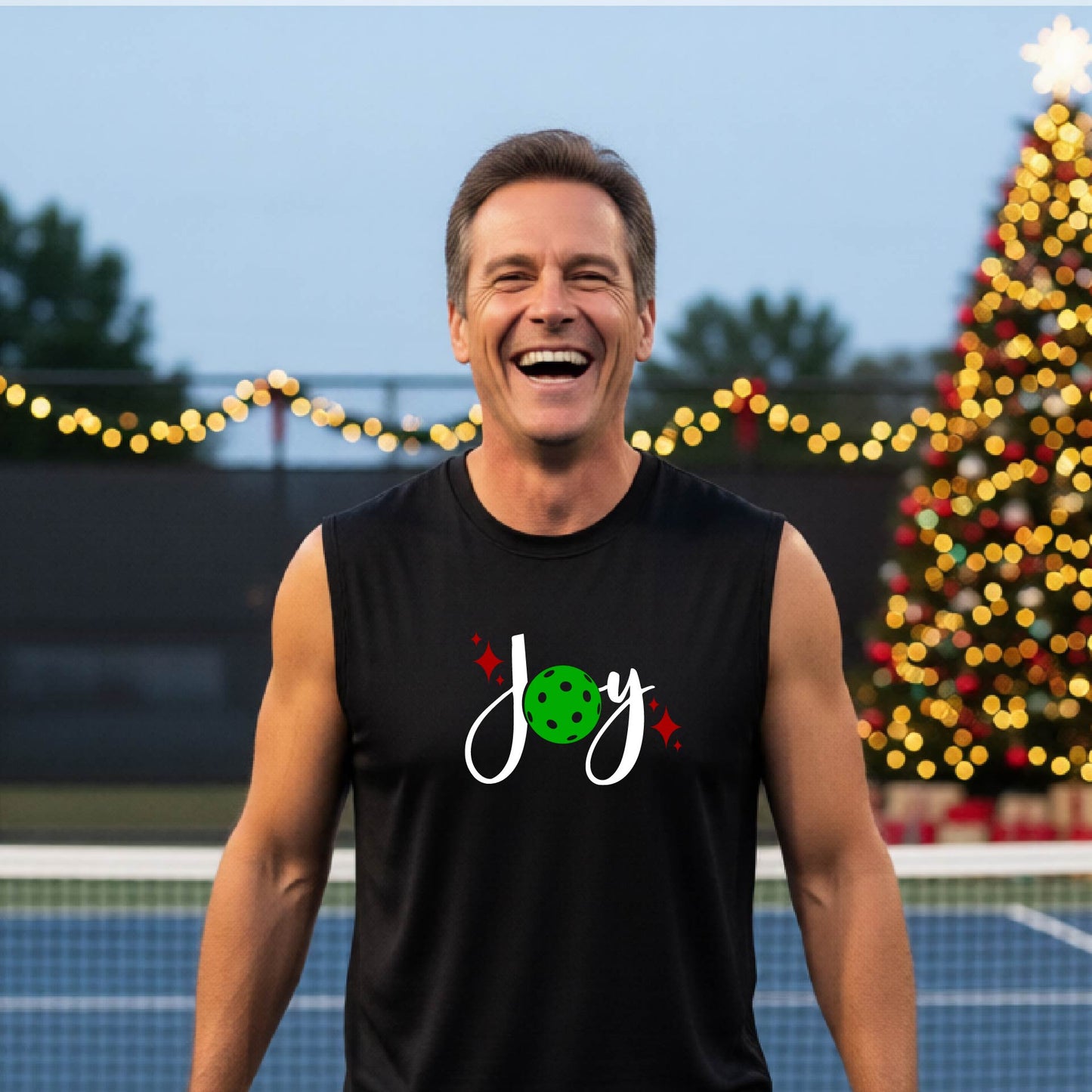 A smiling man wearing a black sleeveless tank top with a 'Joy' pickleball graphic stands on a pickleball court with a decorated Christmas tree and string lights in the background.