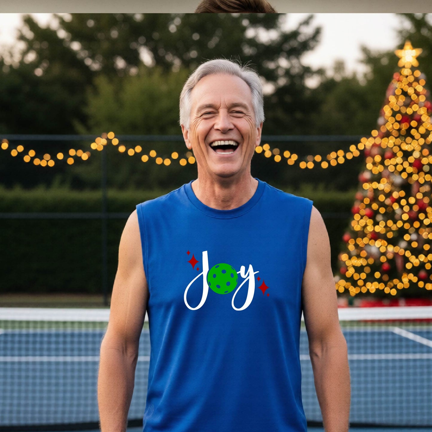A smiling man wearing a royal blue sleeveless tank top with a 'Joy' pickleball graphic stands on a pickleball court with a decorated Christmas tree and string lights in the background.