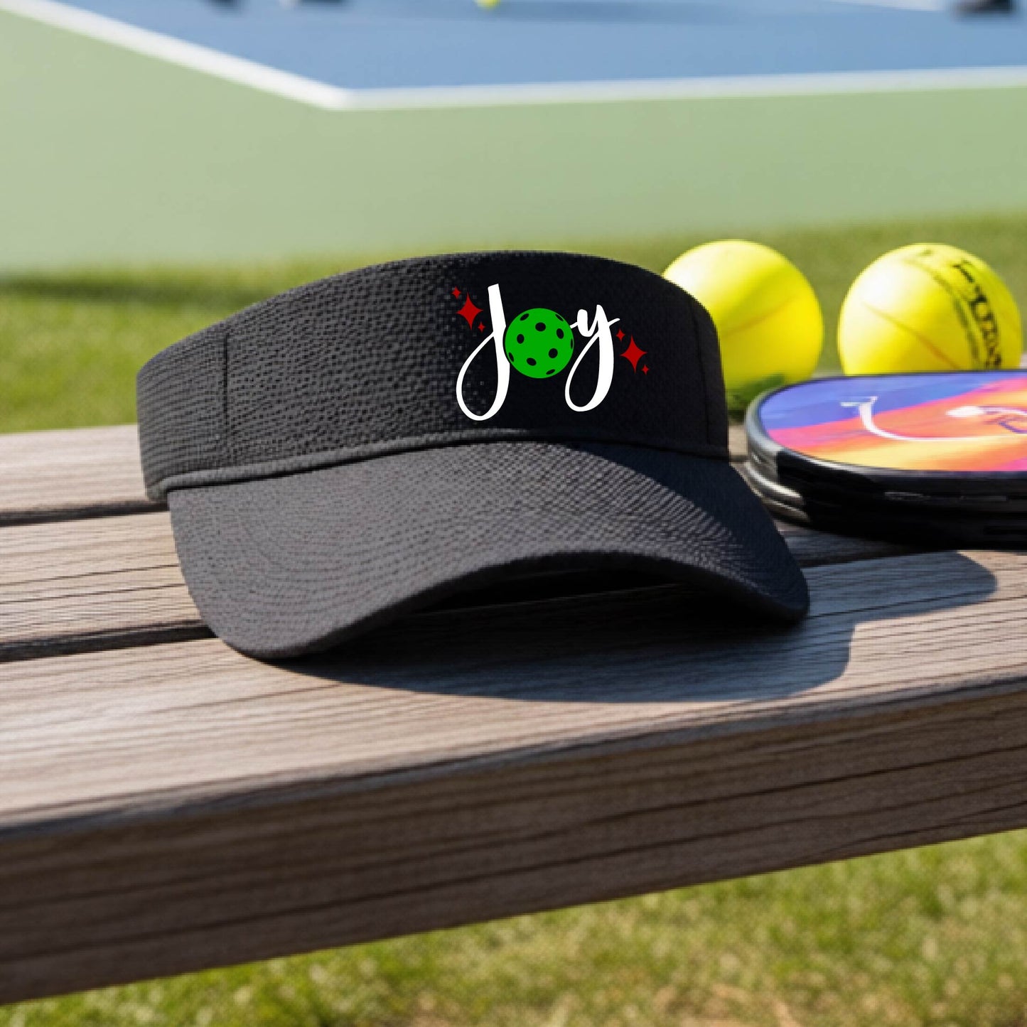 A black visor with a pickleball Christmas graphic of the text "JOY" with the O replaced by a green pickleball. 