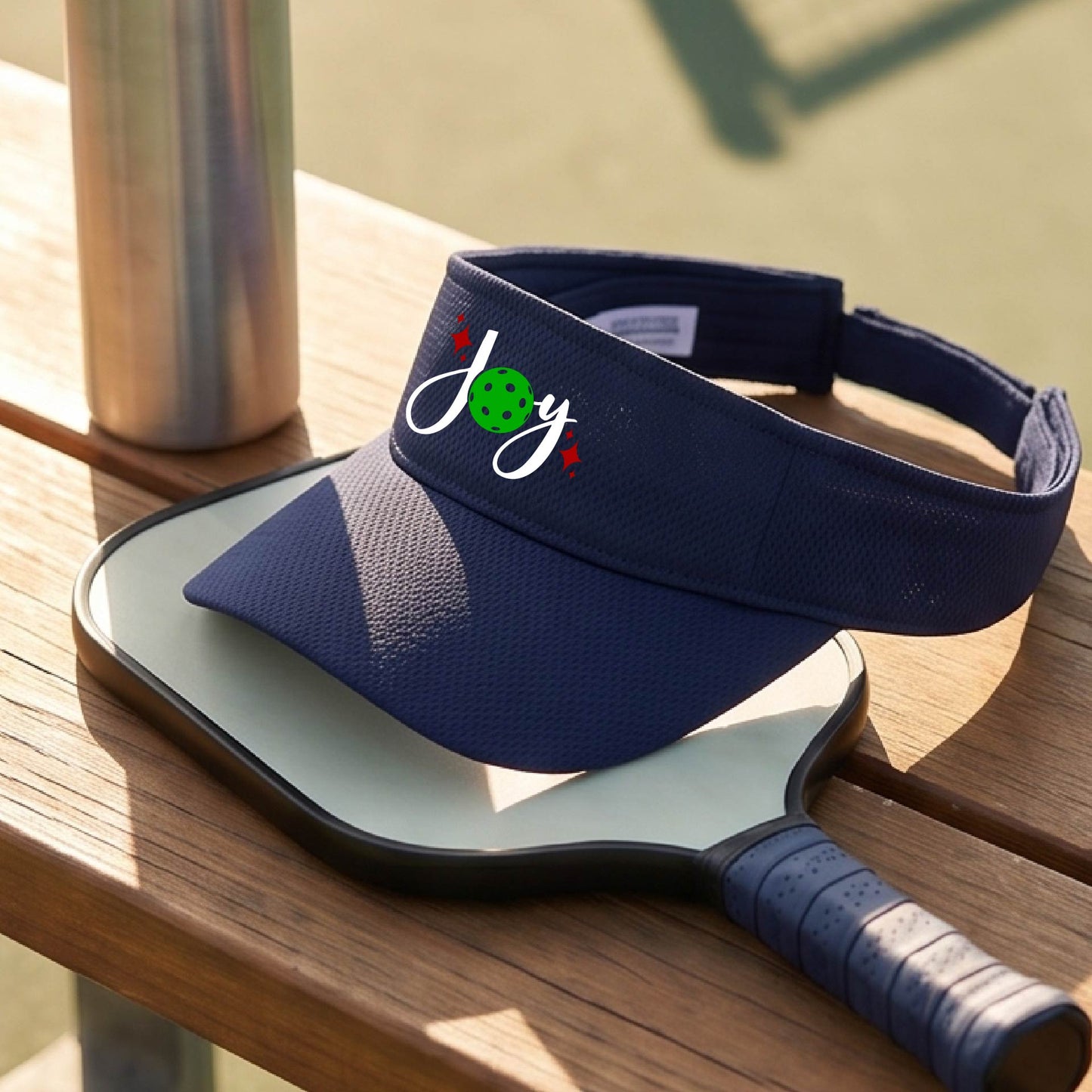 A navy blue visor with a pickleball Christmas graphic of the text "JOY" with the O replaced by a green pickleball. 
