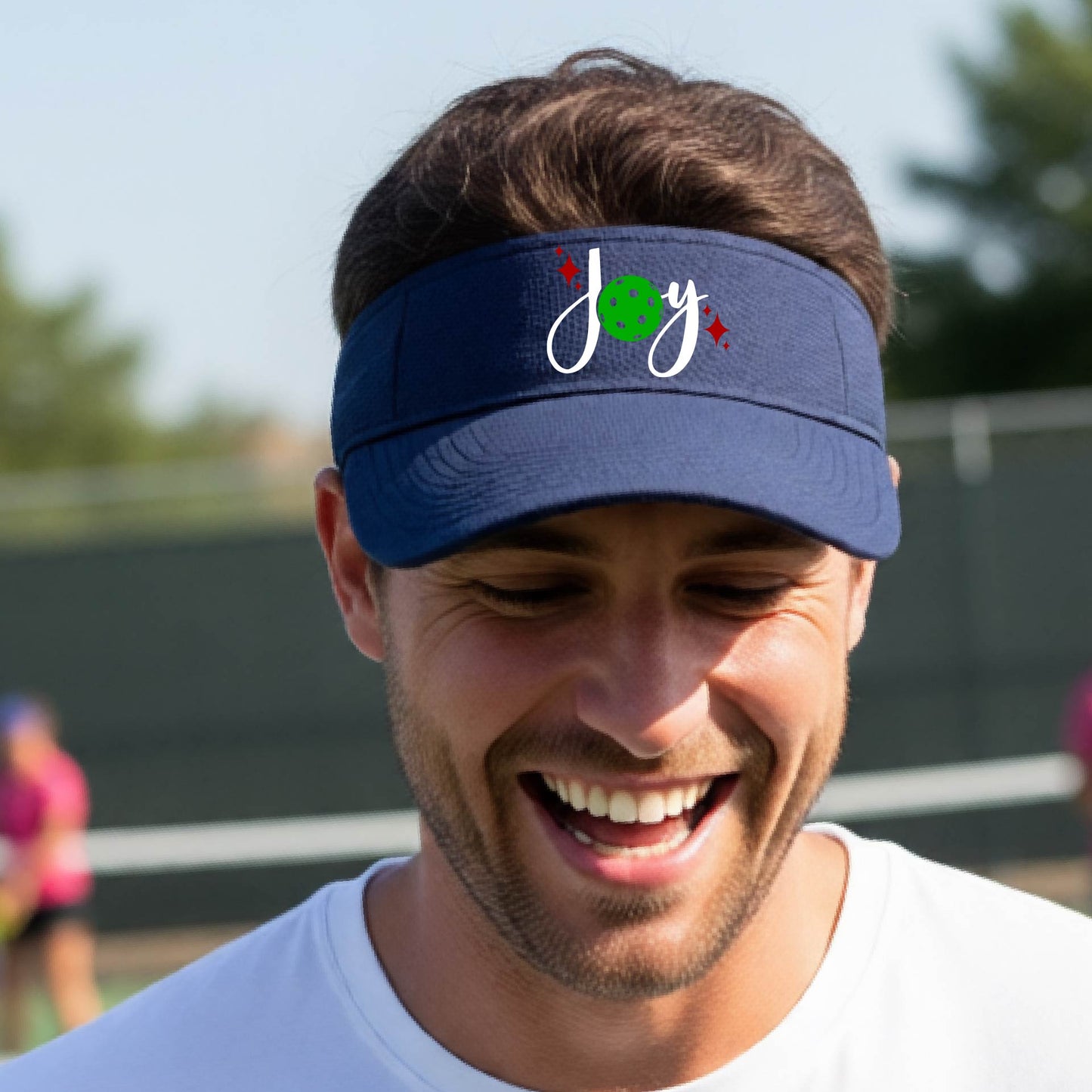 A navy blue visor with a pickleball Christmas graphic of the text "JOY" with the O replaced by a green pickleball. 