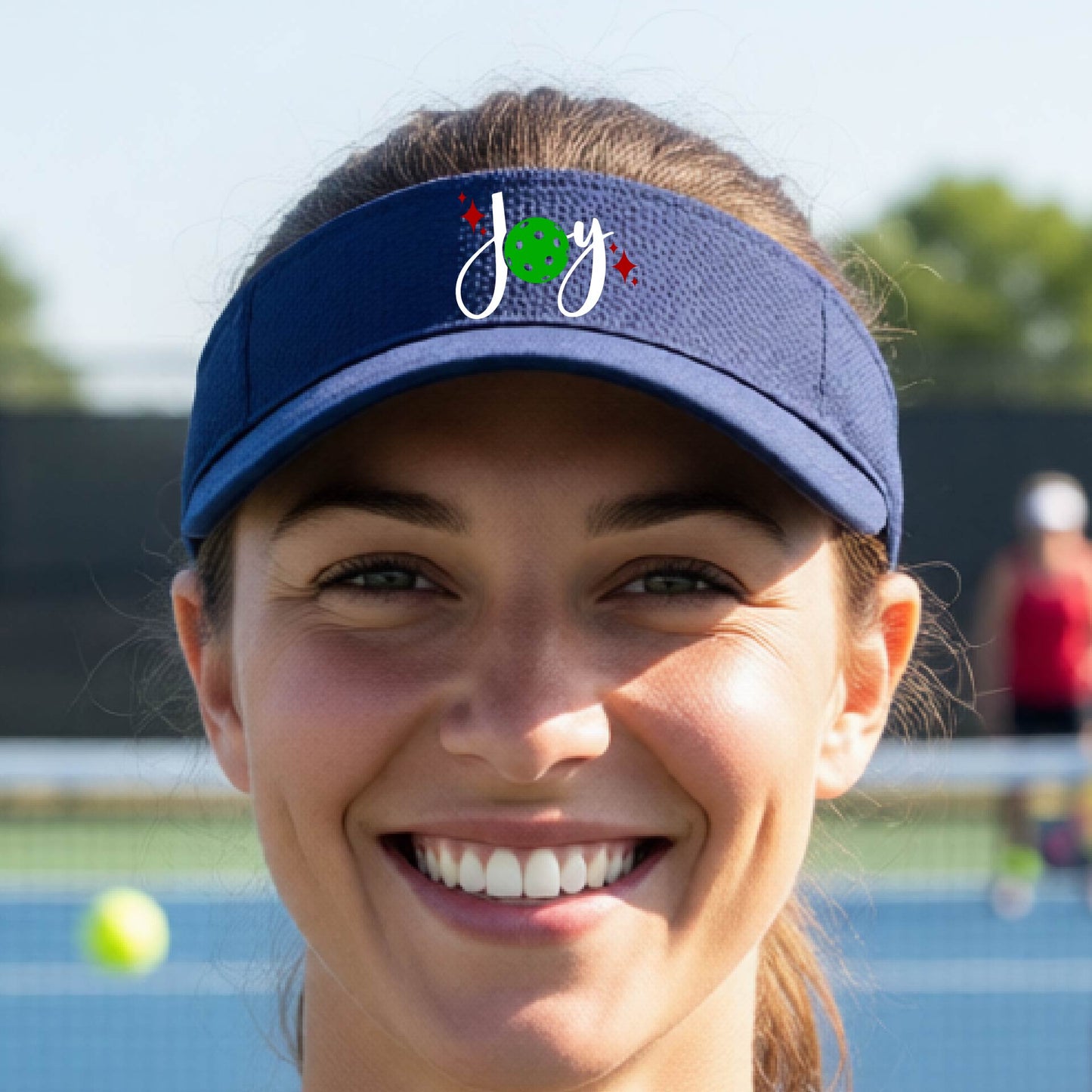 A blue visor with a pickleball Christmas graphic of the text "JOY" with the O replaced by a green pickleball.