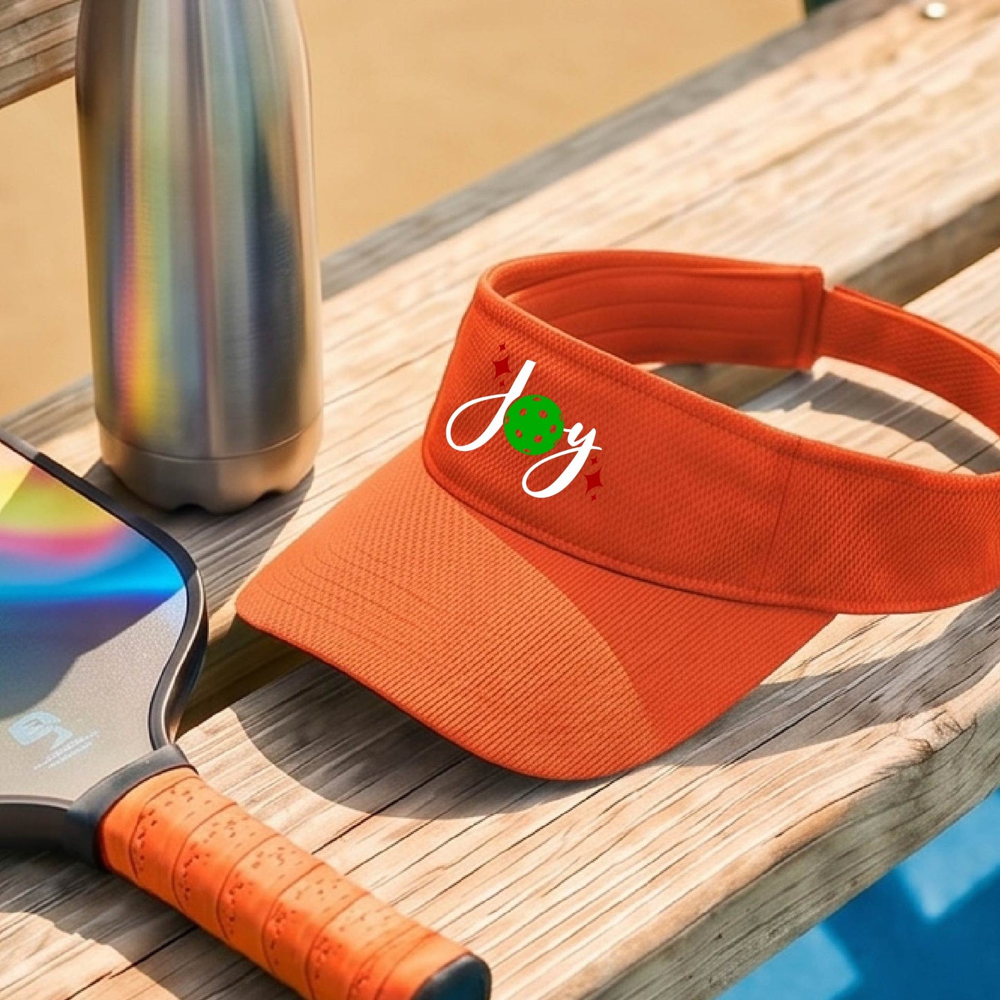 An orange visor with a pickleball Christmas graphic of the text "JOY" with the O replaced by a green pickleball. 