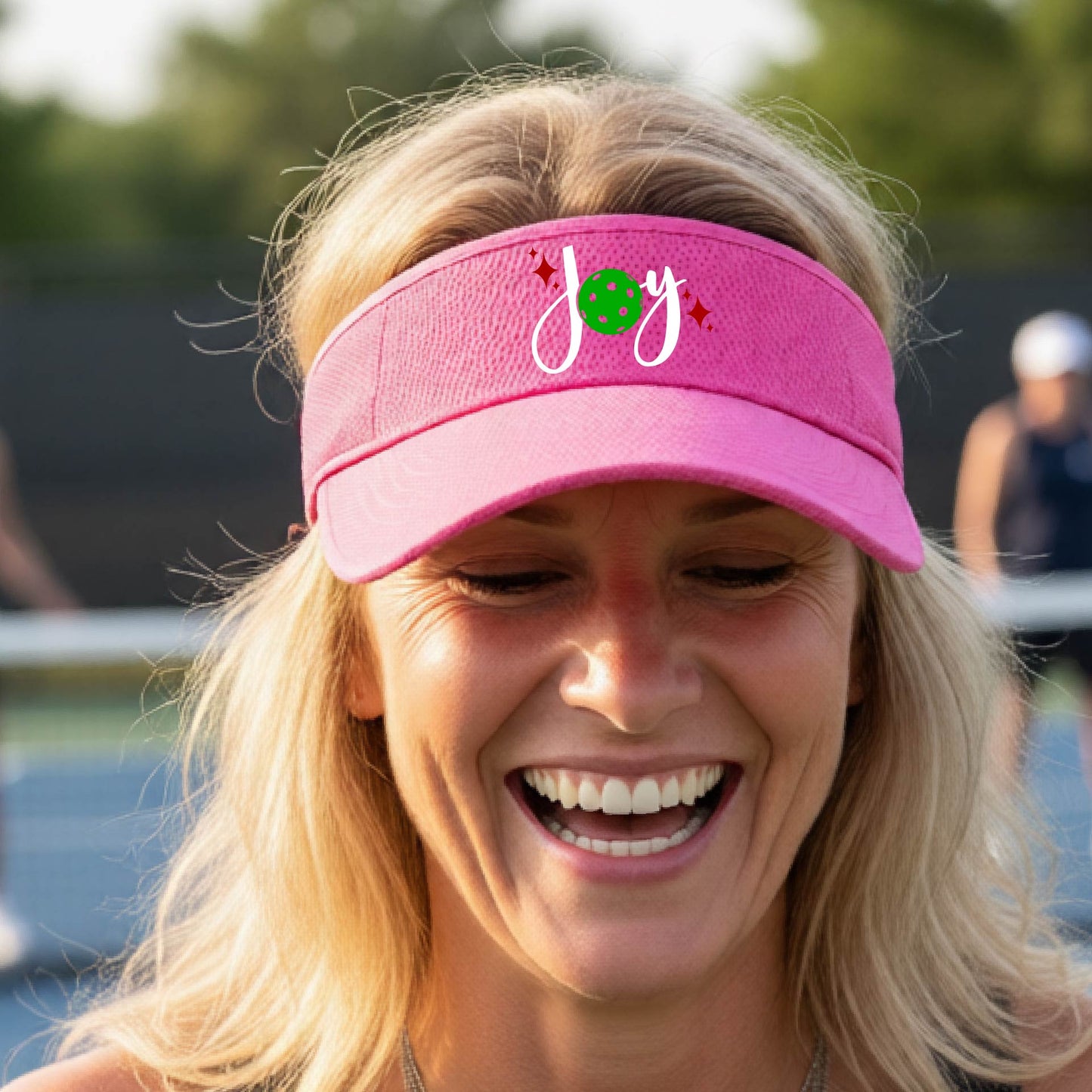 A pink visor with a pickleball Christmas graphic of the text "JOY" with the O replaced by a green pickleball. 