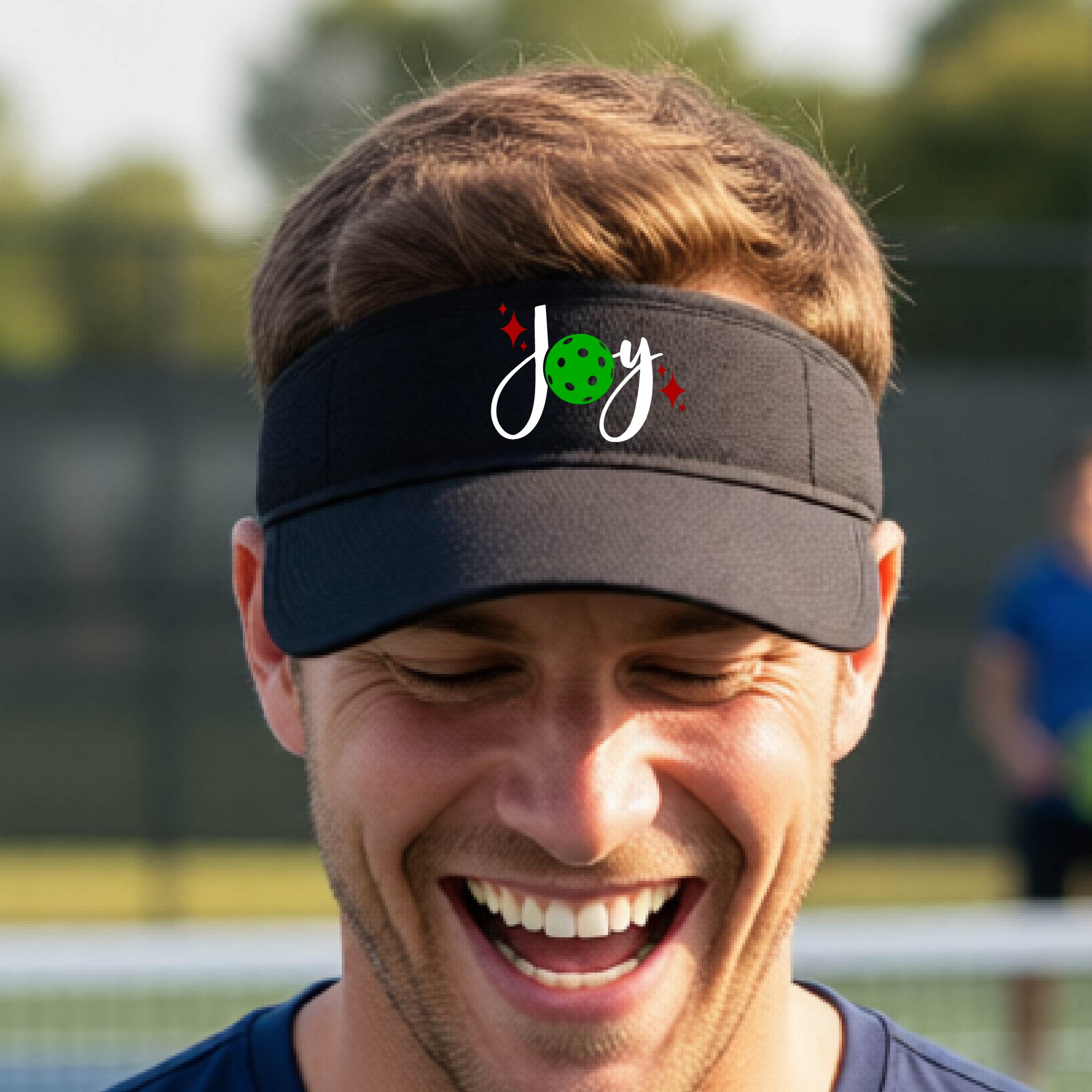 A black visor with a pickleball Christmas graphic of the text "JOY" with the O replaced by a green pickleball. 