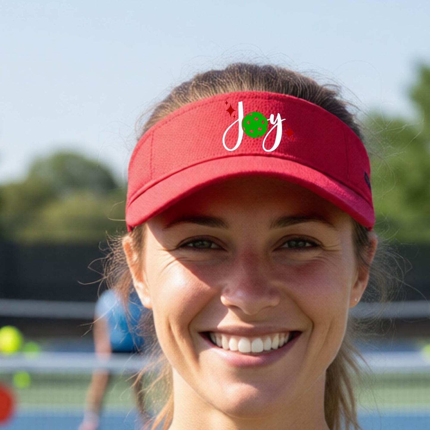 A red visor with a pickleball Christmas graphic of the text "JOY" with the O replaced by a green pickleball. 