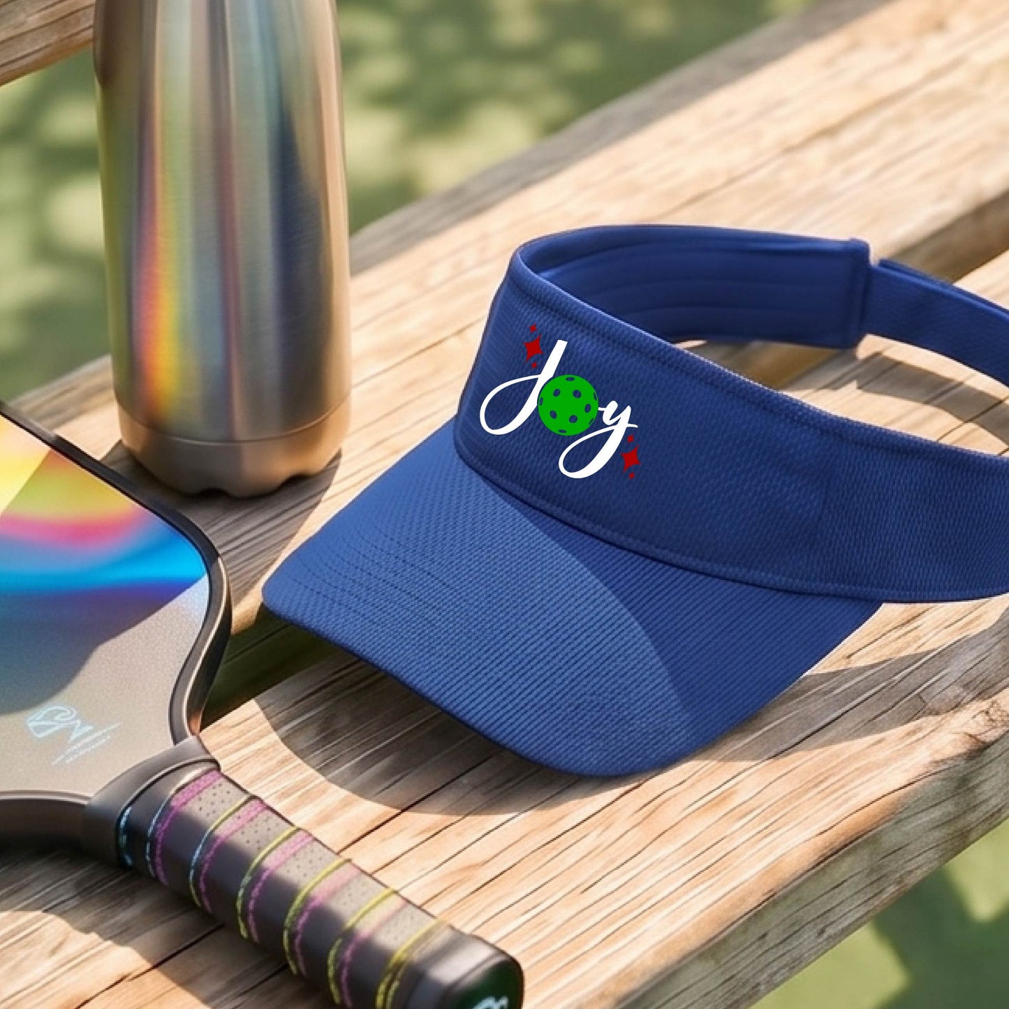 A royal blue visor with a pickleball Christmas graphic of the text "JOY" with the O replaced by a green pickleball. 