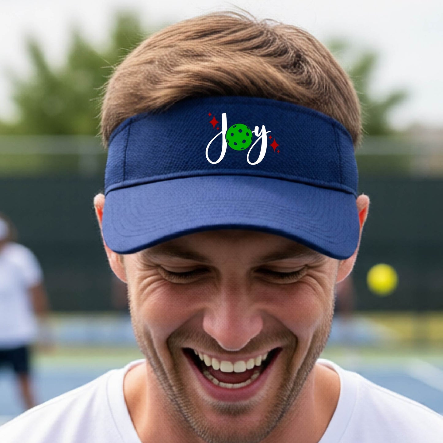 A royal blue visor with a pickleball Christmas graphic of the text "JOY" with the O replaced by a green pickleball. 