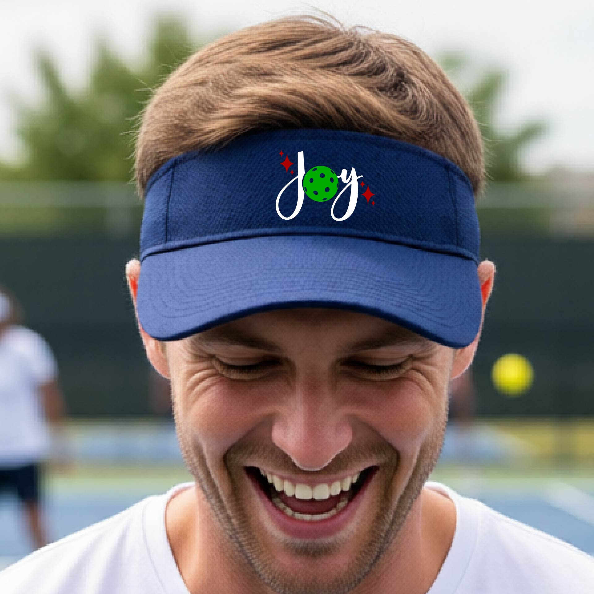 A royal blue visor with a pickleball Christmas graphic of the text "JOY" with the O replaced by a green pickleball. 