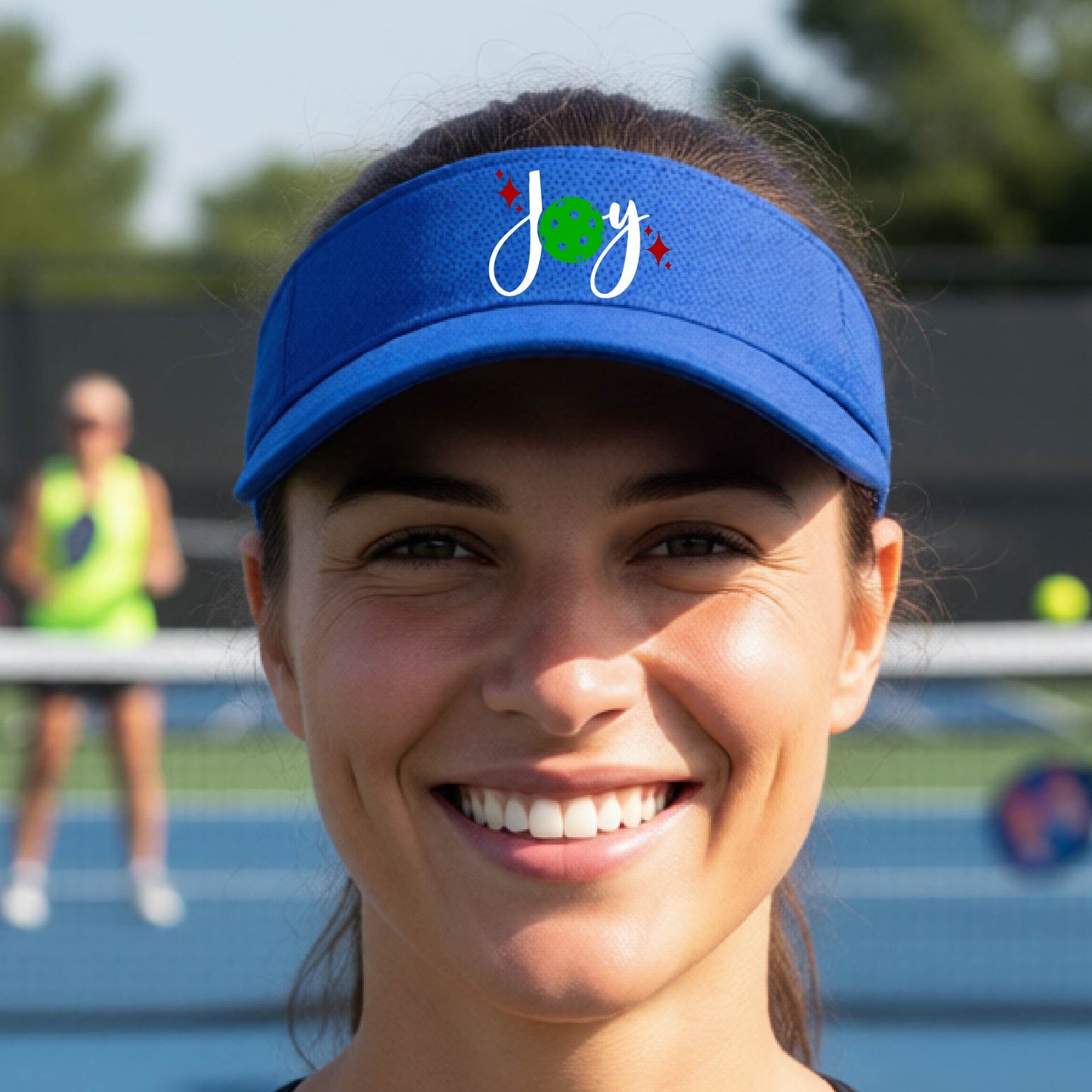 A royal blue visor with a pickleball Christmas graphic of the text "JOY" with the O replaced by a green pickleball. 