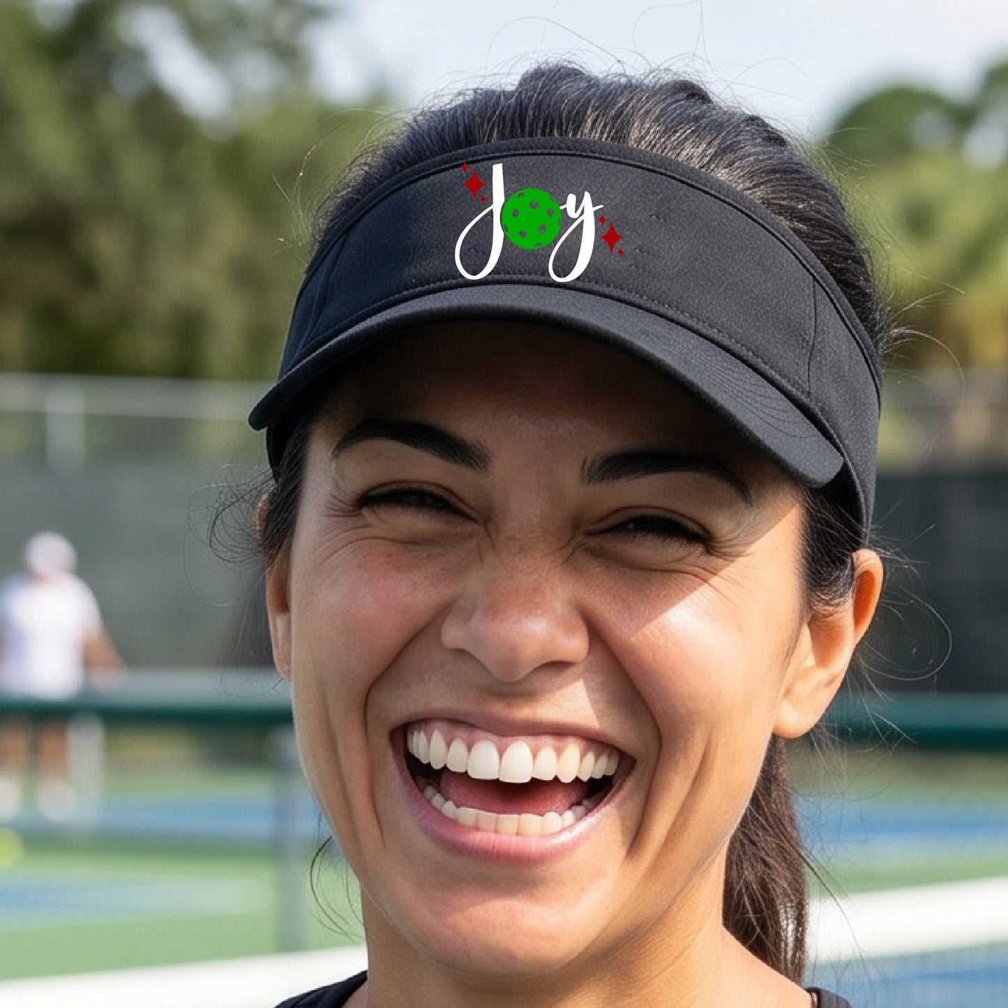 A black visor with a pickleball Christmas graphic of the text "JOY" with the O replaced by a green pickleball. 