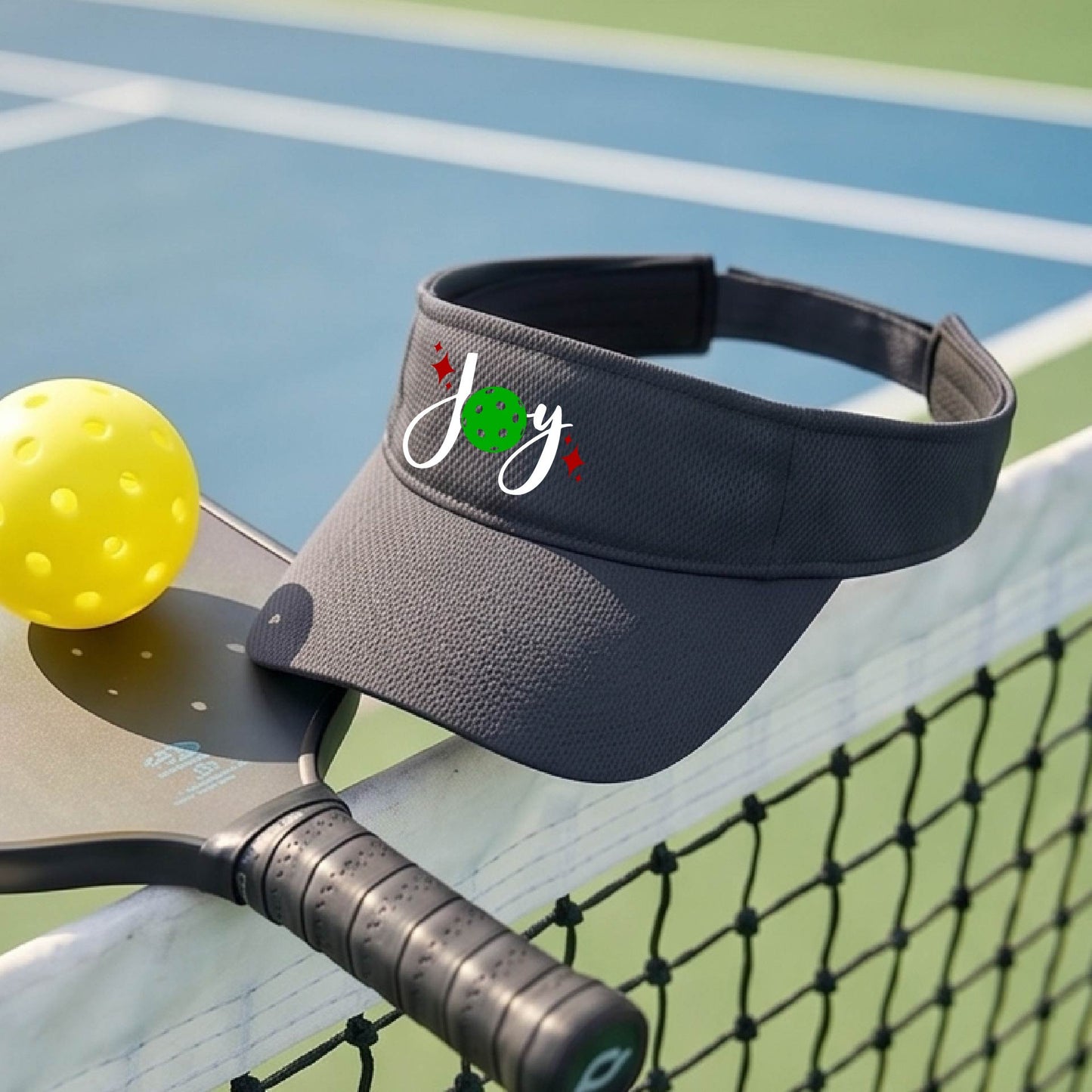 A graphite gray visor with a pickleball Christmas graphic of the text "JOY" with the O replaced by a green pickleball. 