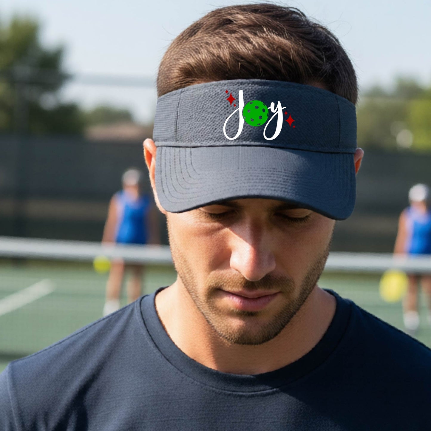 A graphite gray visor with a pickleball Christmas graphic of the text "JOY" with the O replaced by a green pickleball. 