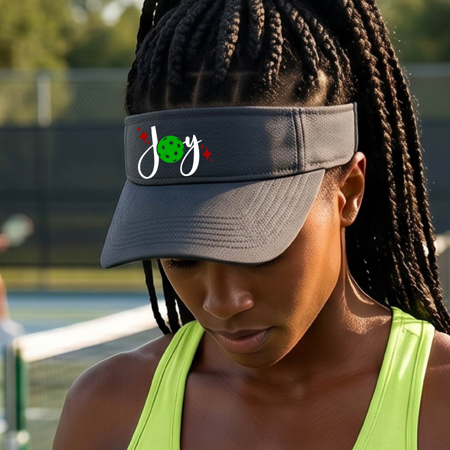 A graphite gray visor with a pickleball Christmas graphic of the text "JOY" with the O replaced by a green pickleball. 