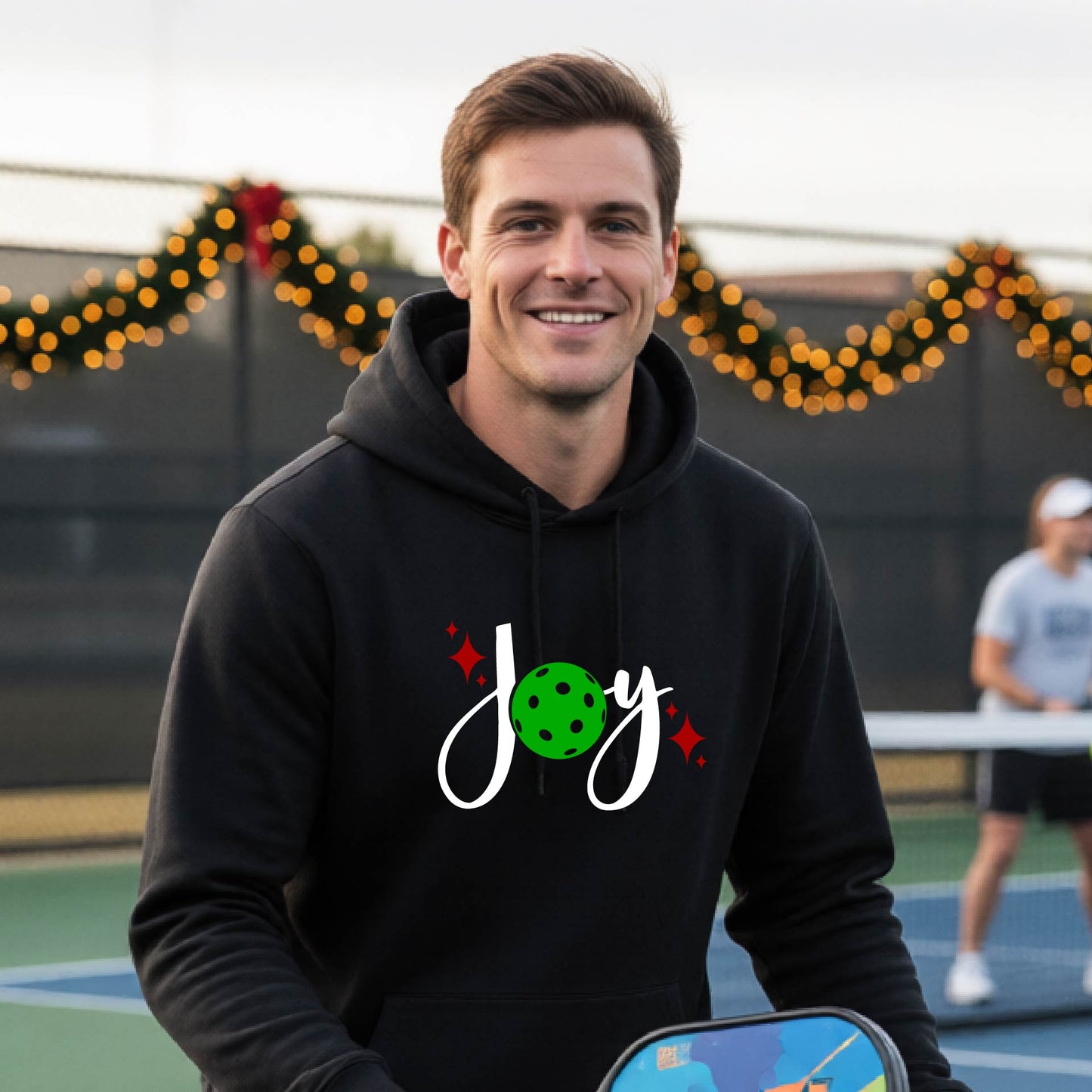 A smiling man wearing a black hoodie with a 'Joy' graphic on the chest stands on an outdoor pickleball court. The graphic features the word 'Joy' in white script with a green pickleball 'o' and small red star accents.