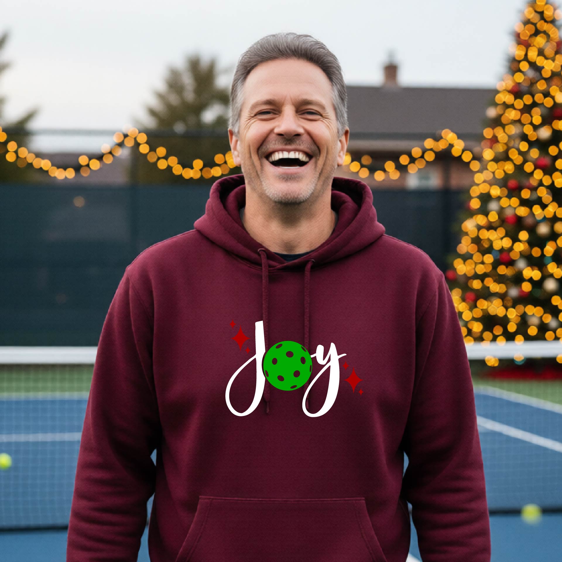 A smiling man wearing a maroon hoodie with a 'Joy' graphic on the chest stands on an outdoor pickleball court. The graphic features the word 'Joy' in white script with a green pickleball 'o' and small red star accents.