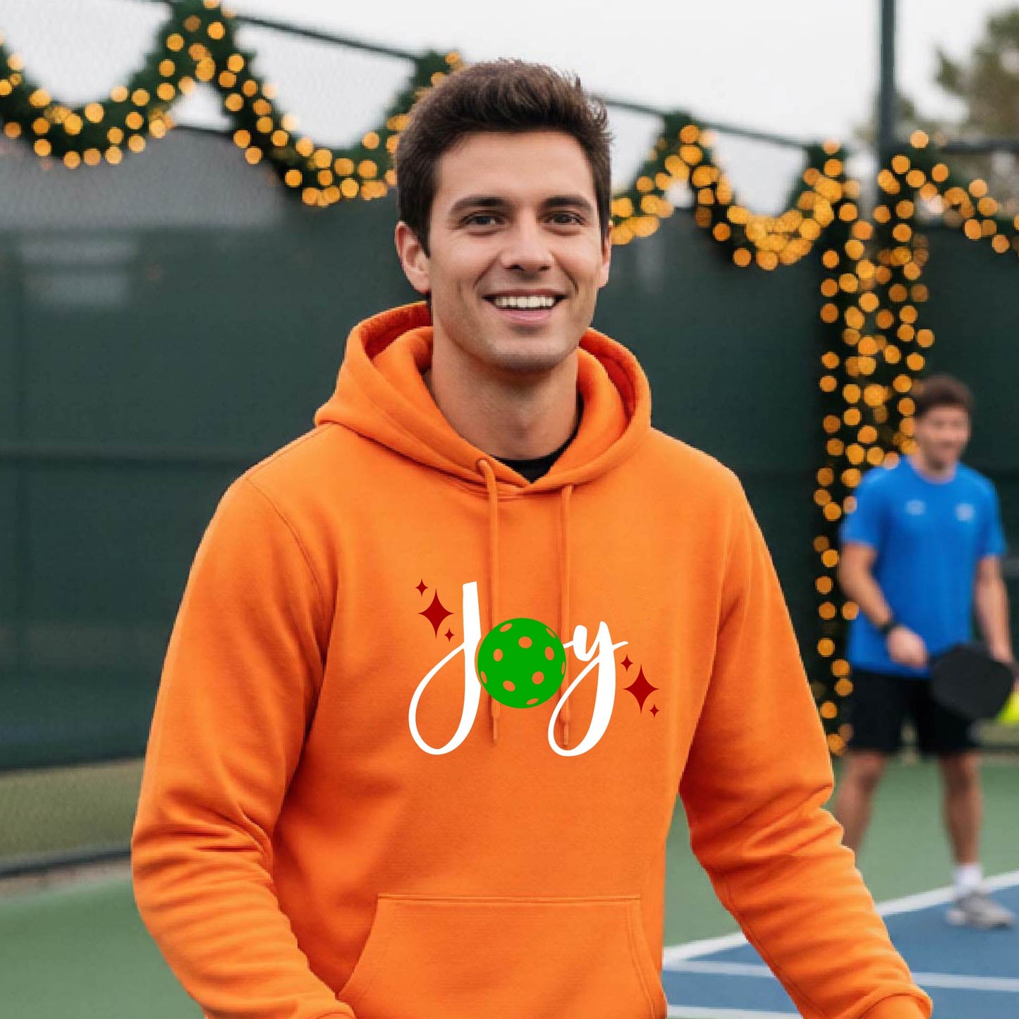 A smiling man wearing a bright orange hoodie with a 'Joy' graphic on the chest stands on an outdoor pickleball court. The graphic features the word 'Joy' in white script with a green pickleball 'o' and small red star accents.