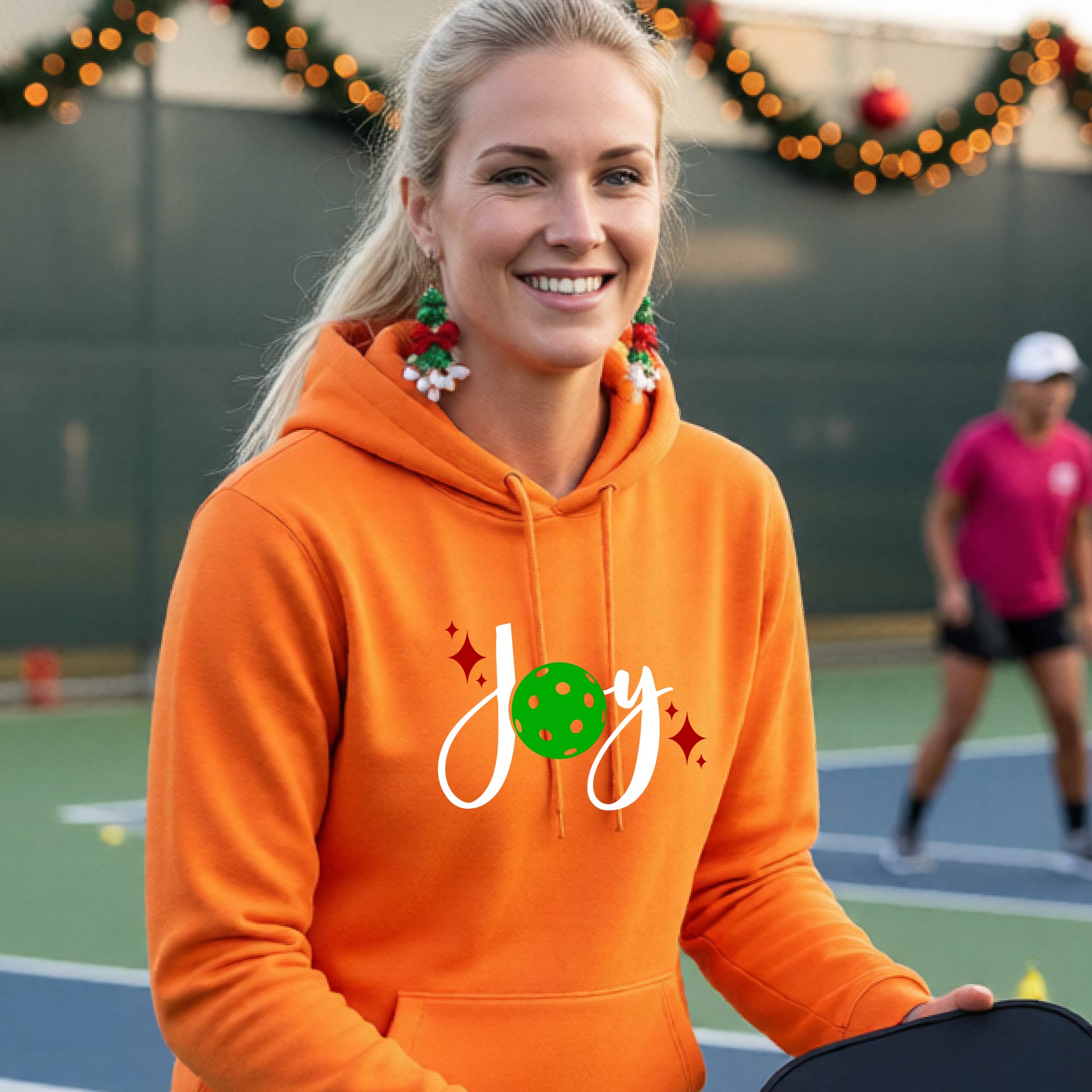 A smiling woman wearing a bright orange hoodie with a 'Joy' graphic on the chest stands on an outdoor pickleball court. The graphic features the word 'Joy' in white script with a green pickleball 'o' and small red star accents.
