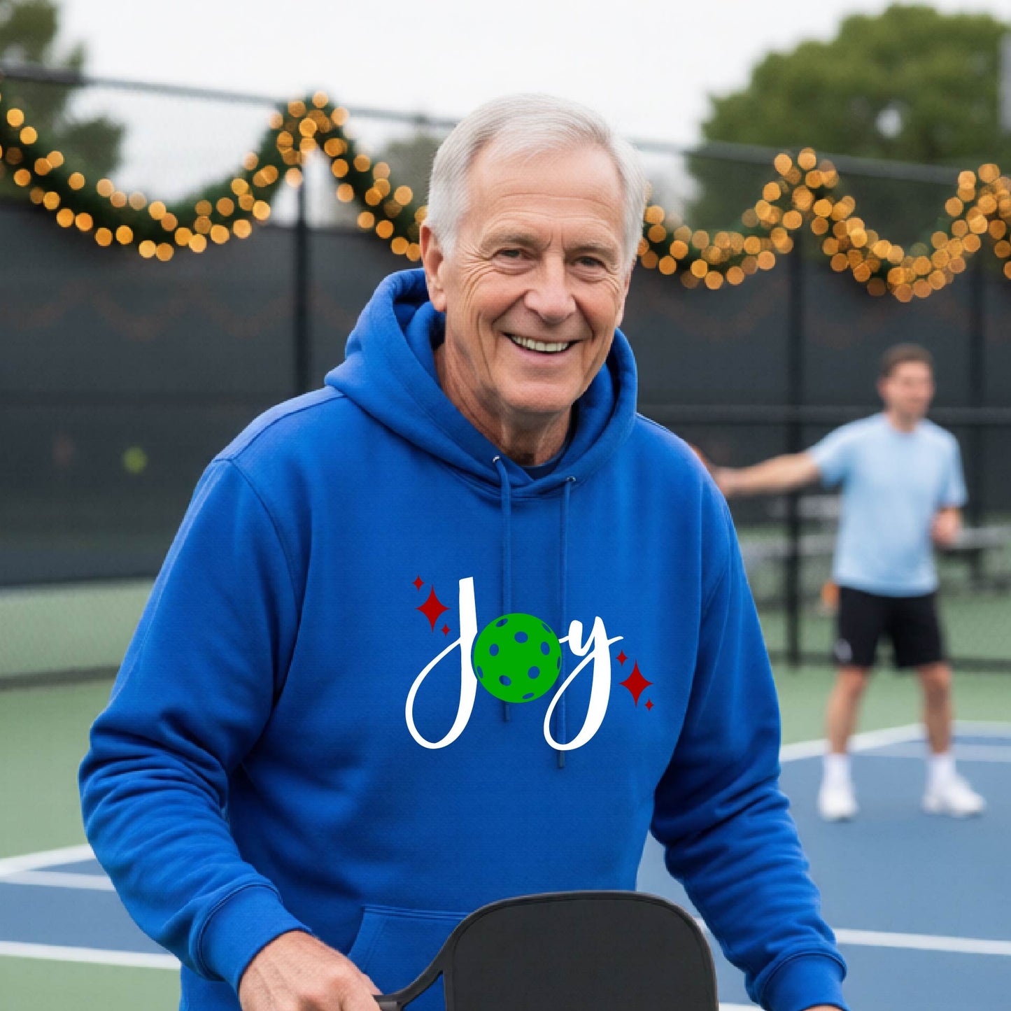 A smiling man wearing a royal blue hoodie with a 'Joy' graphic on the chest stands on an outdoor pickleball court. The graphic features the word 'Joy' in white script with a green pickleball 'o' and small red star accents.