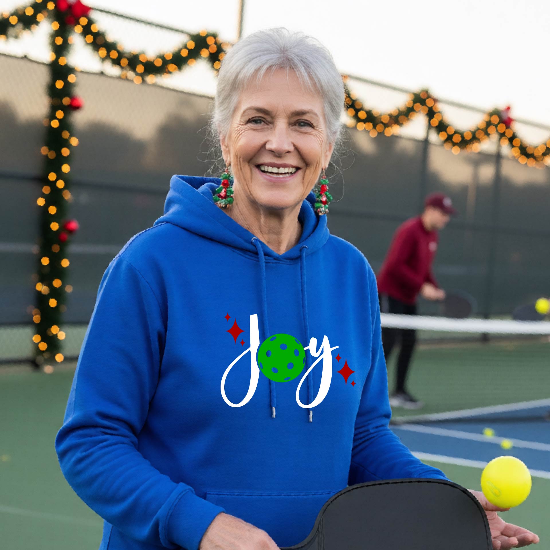 A smiling woman wearing a royal blue hoodie with a 'Joy' graphic on the chest stands on an outdoor pickleball court. The graphic features the word 'Joy' in white script with a green pickleball 'o' and small red star accents.