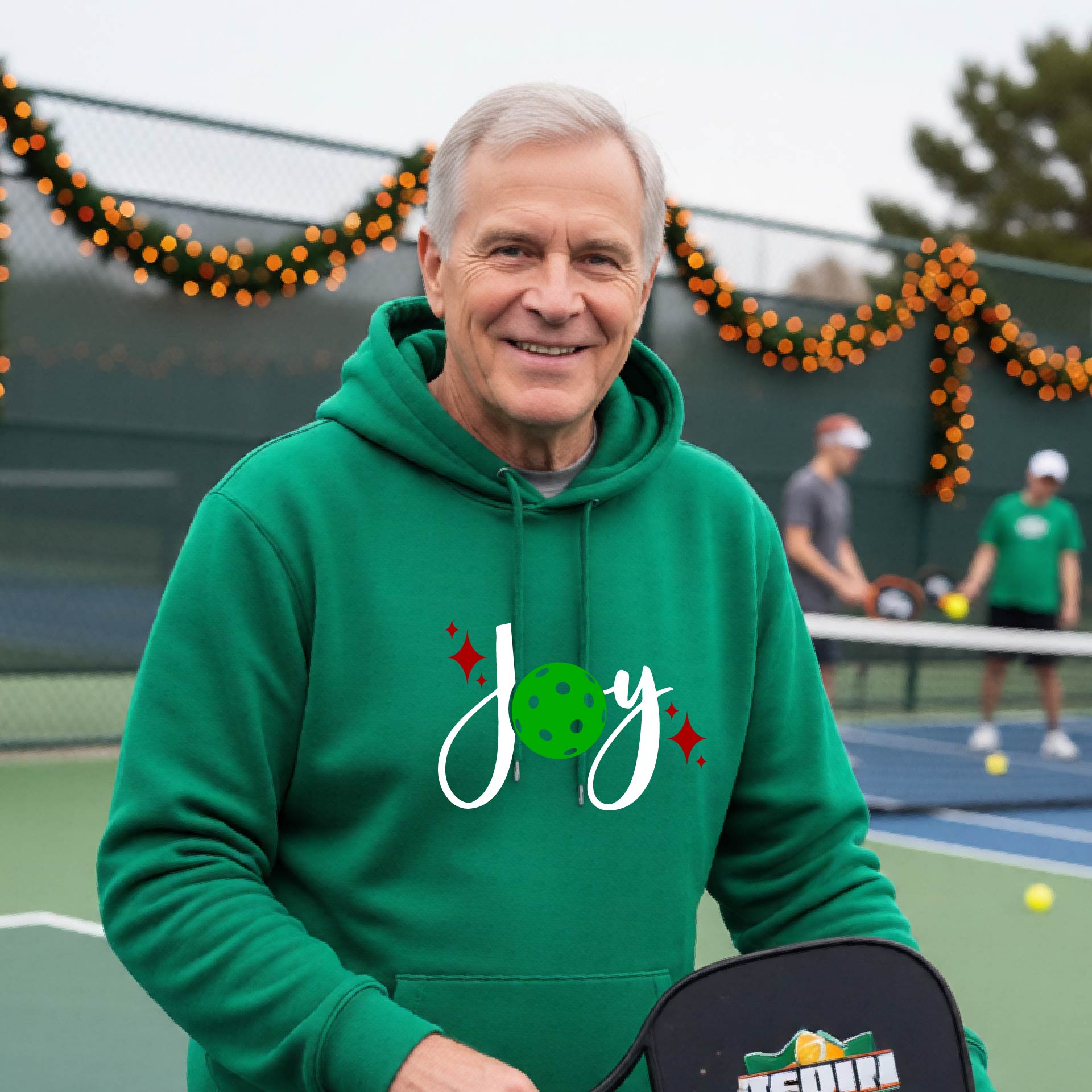 A smiling man wearing a green hoodie with a 'Joy' graphic on the chest stands on an outdoor pickleball court. The graphic features the word 'Joy' in white script with a green pickleball 'o' and small red star accents.