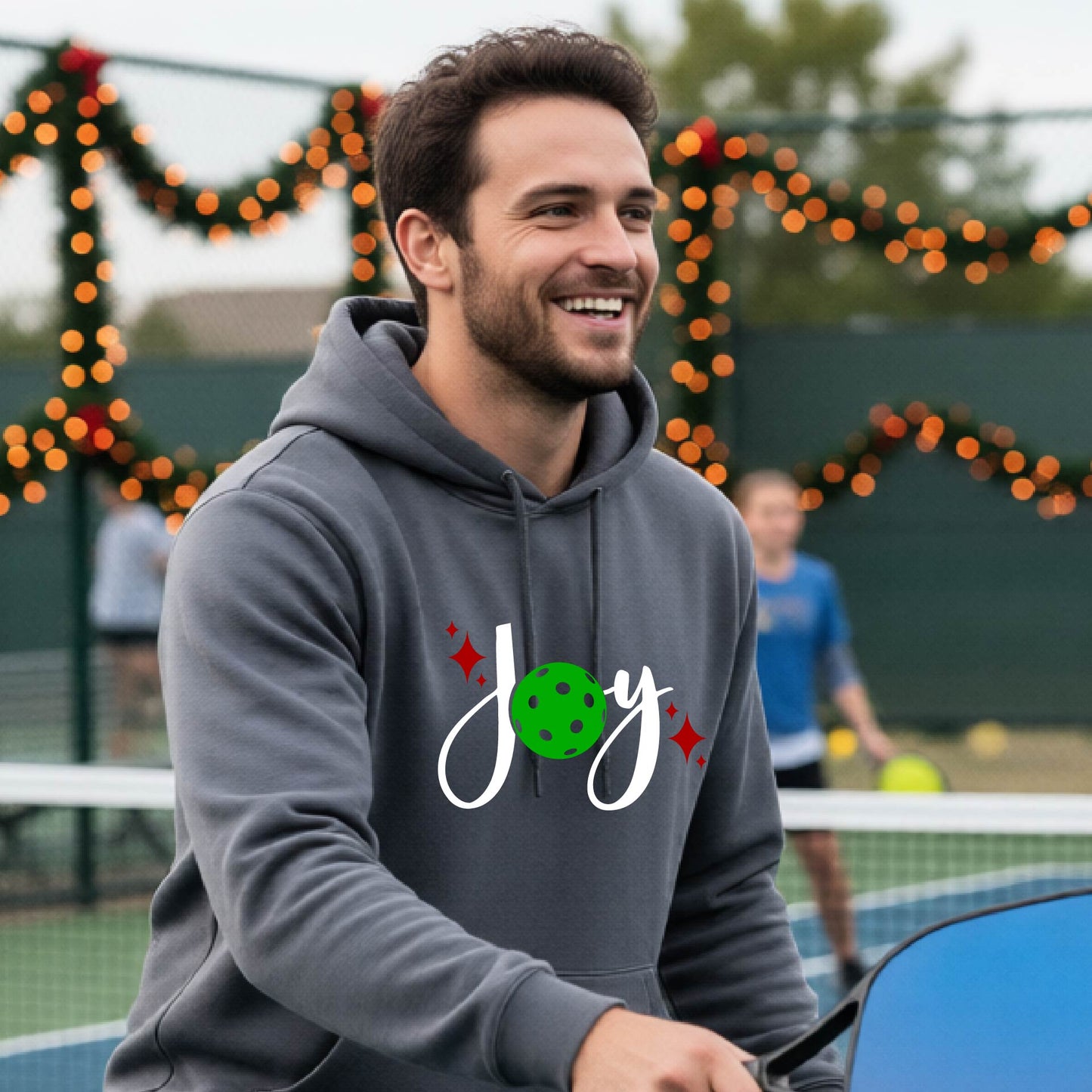 A smiling man wearing a gray hoodie with a 'Joy' graphic on the chest stands on an outdoor pickleball court. The graphic features the word 'Joy' in white script with a green pickleball 'o' and small red star accents.