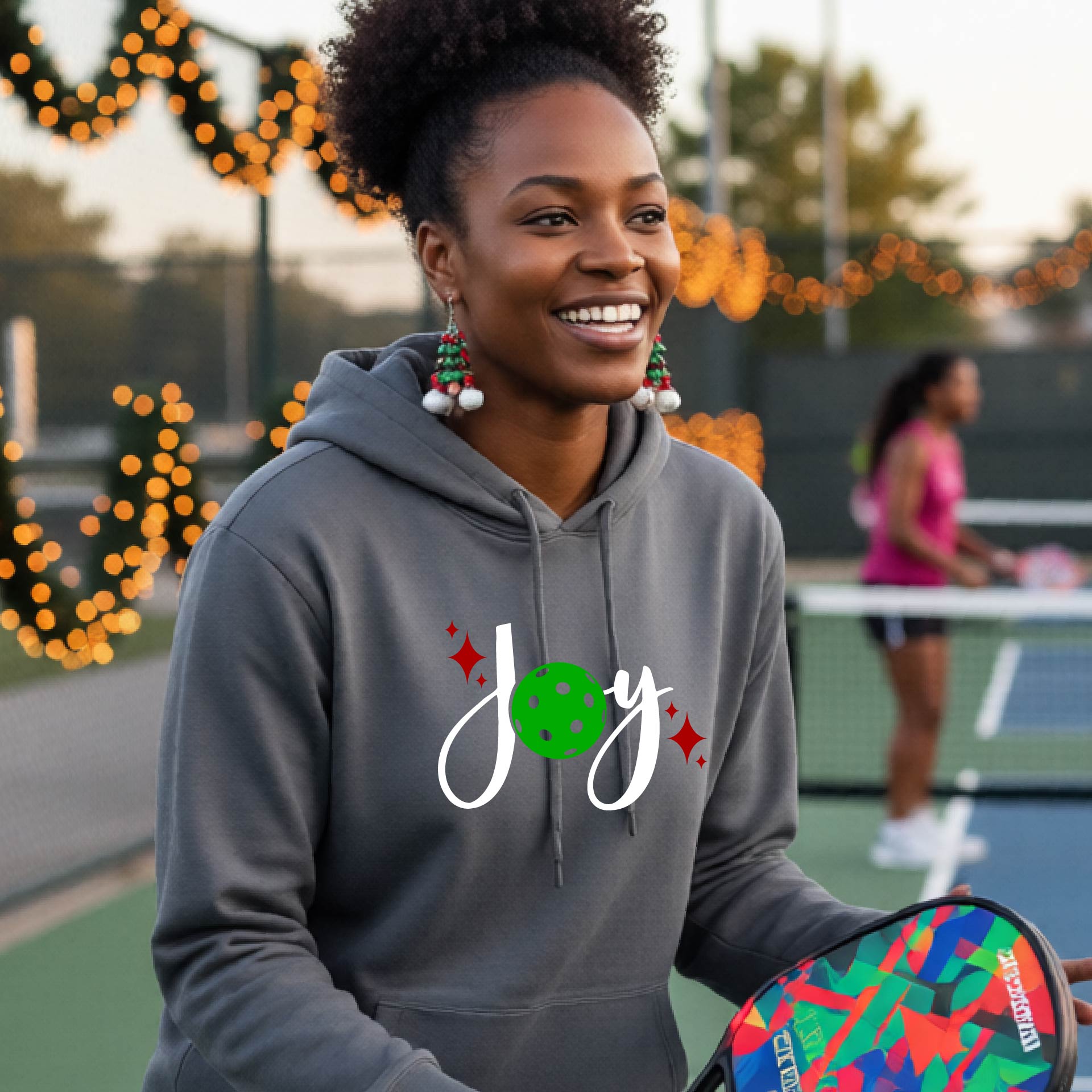 woman wearing a grey hoodie with a 'Joy' graphic on the chest stands on an outdoor pickleball court. The graphic features the word 'Joy' in white script with a green pickleball 'o' and small red star accents.