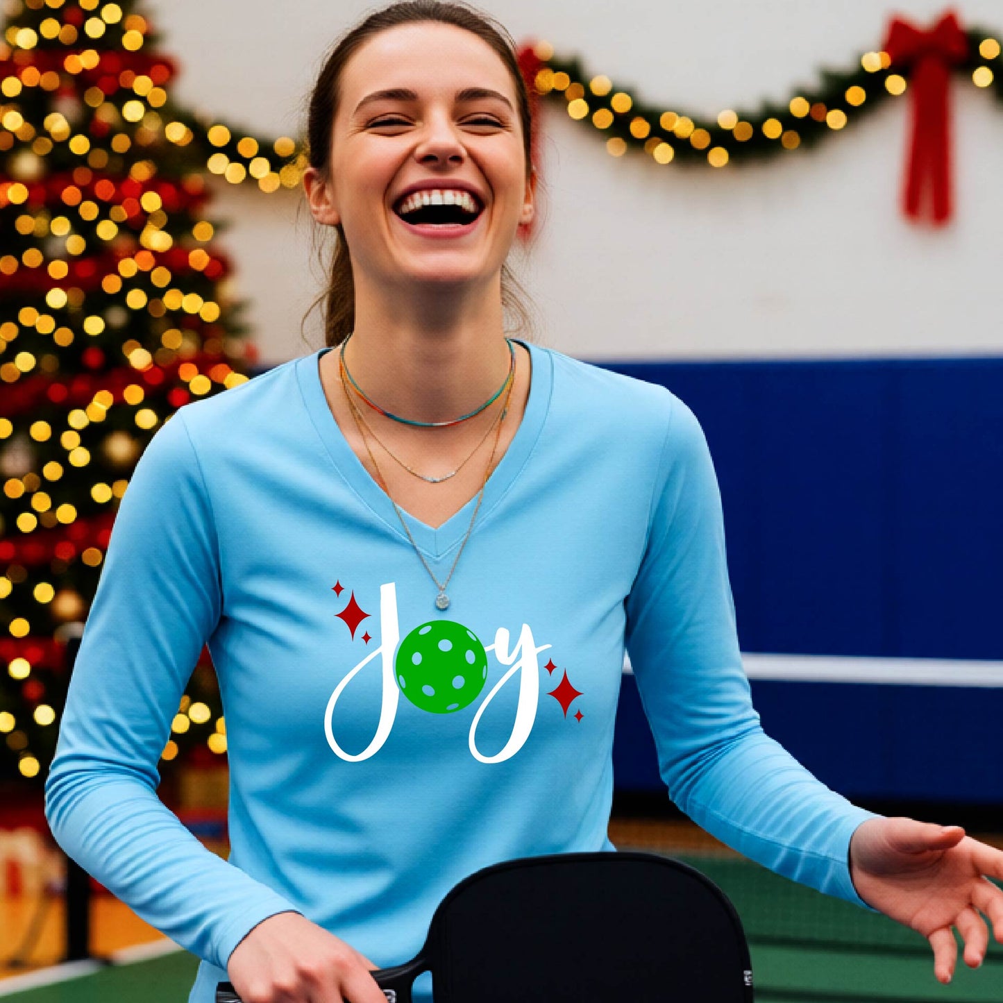 A women's light blue long-sleeve v-neck shirt featuring a pickleball graphic with text "JOY"  with the O replaced by a green pickleball and  Christmas themed background.