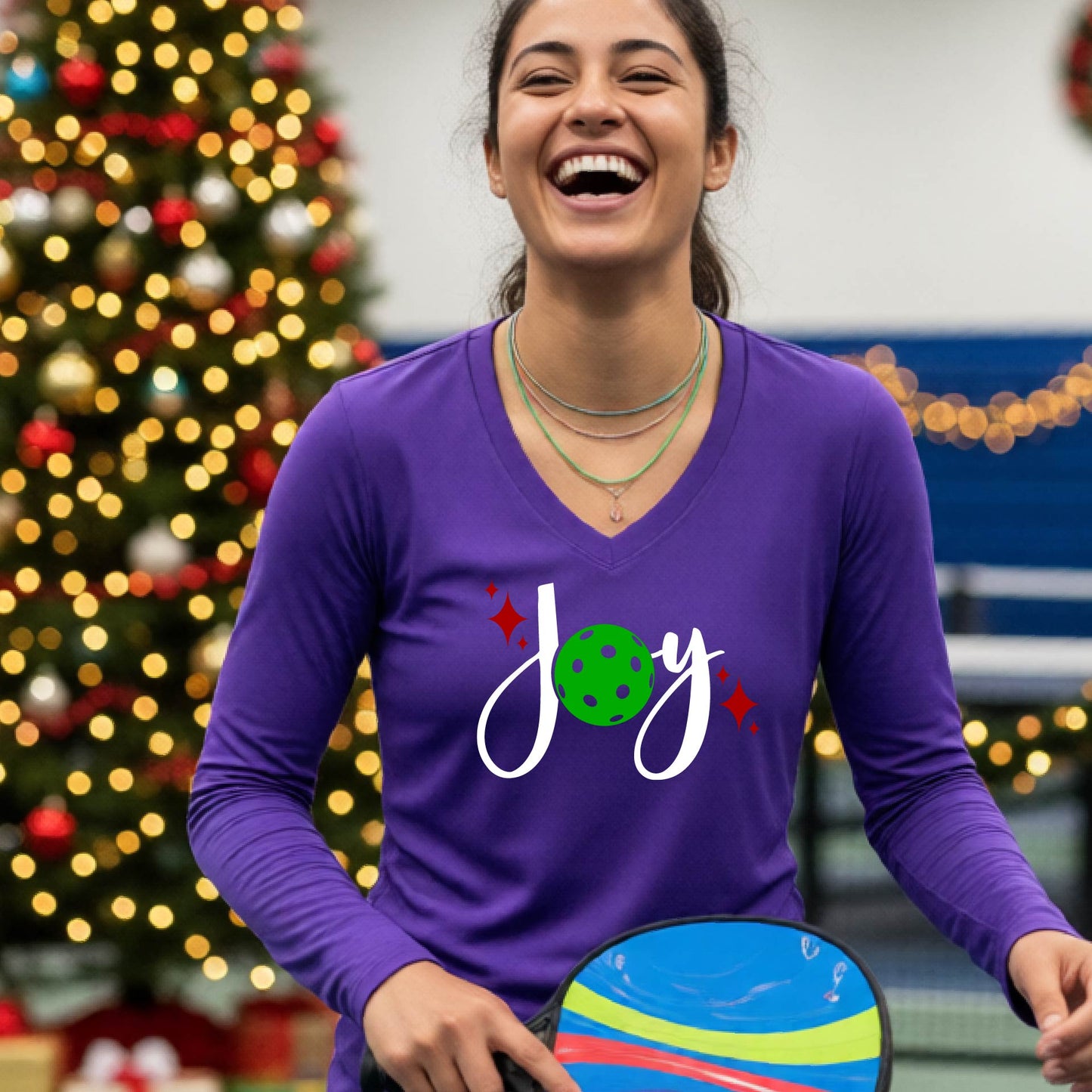 A women's purple long-sleeve v-neck shirt featuring a pickleball graphic with text "JOY"  with the O replaced by a green pickleball and  Christmas themed background.