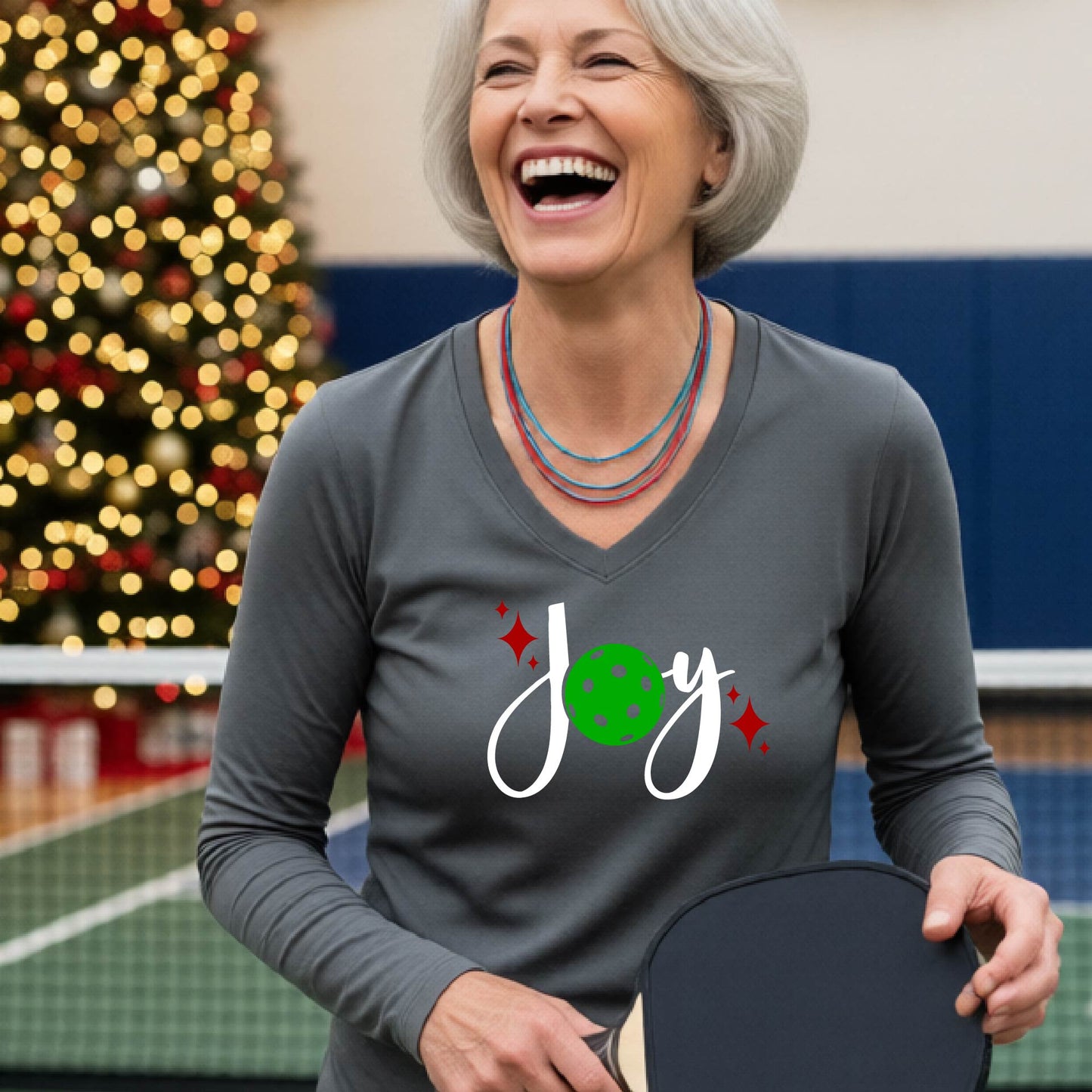 A women's gray long-sleeve v-neck shirt featuring a pickleball graphic with text "JOY"  with the O replaced by a green pickleball and  Christmas themed background.