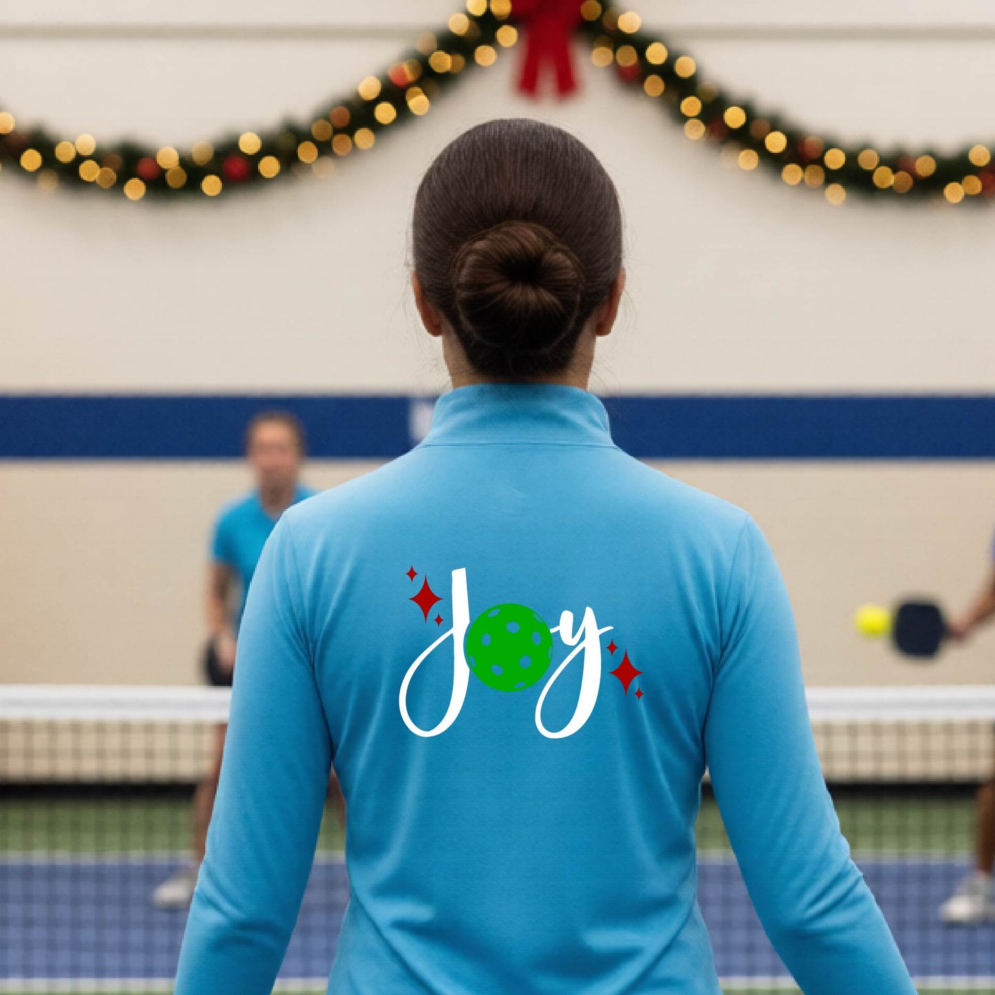 A woman viewed from behind on an indoor pickleball court, wearing a light blue long-sleeve shirt with a 'Joy' graphic on the back. The graphic features a white script 'J' and 'o', a green pickleball, and small red sparkles.