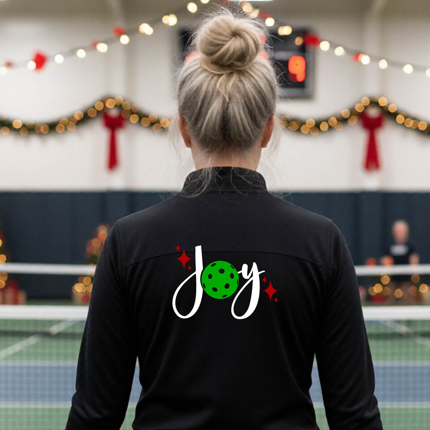 A woman viewed from behind on an indoor pickleball court, wearing a black long-sleeve shirt with a 'Joy' graphic on the back. The graphic features a white script 'J' and 'o', a green pickleball, and small red sparkles.
