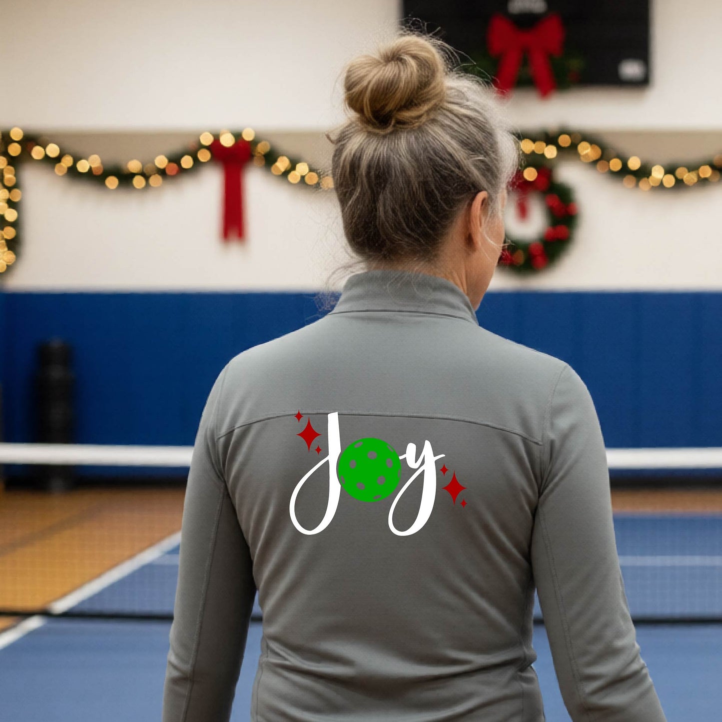 A woman viewed from behind on an indoor pickleball court, wearing a gray long-sleeve shirt with a 'Joy' graphic on the back. The graphic features a white script 'J' and 'o', a green pickleball, and small red sparkles.