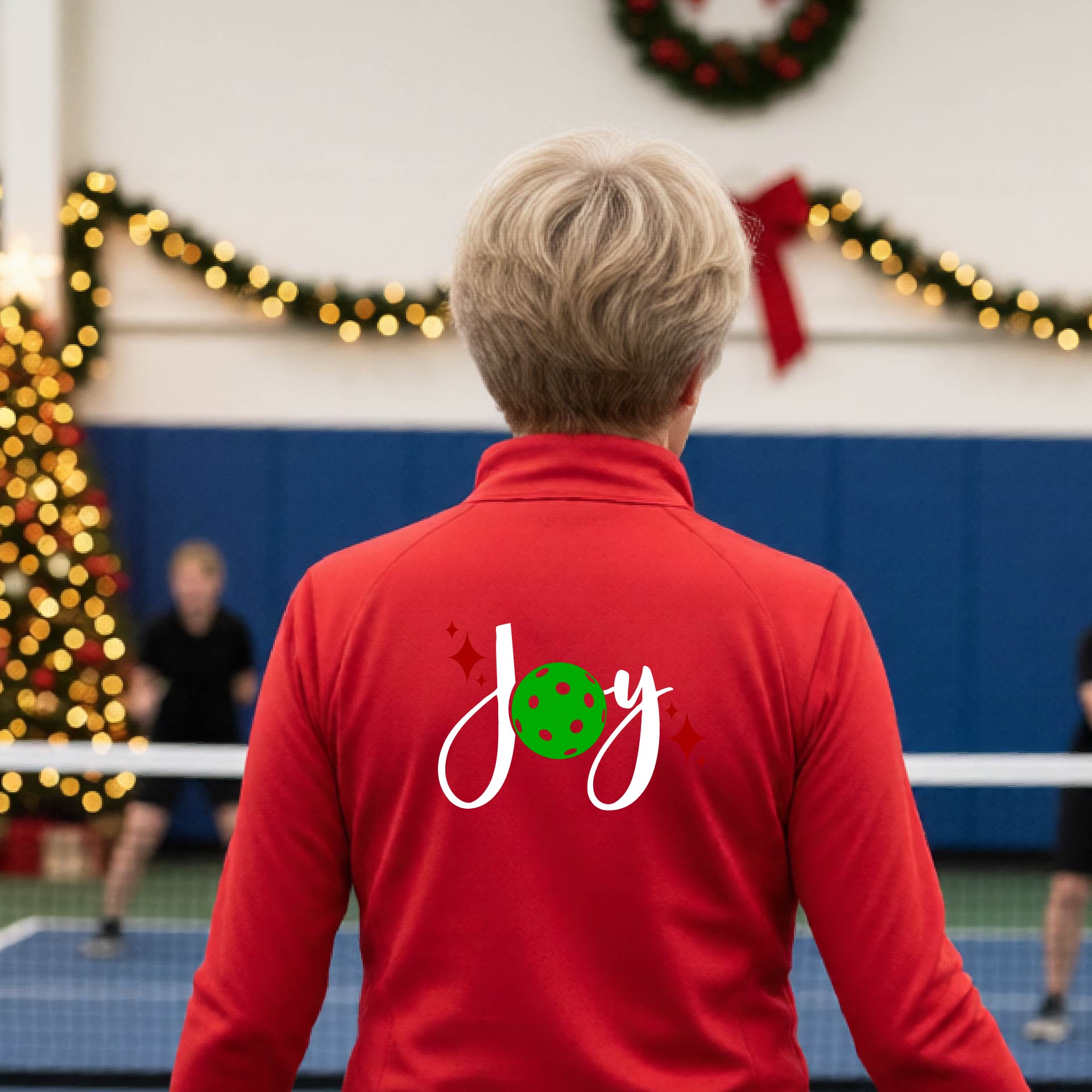 A woman viewed from behind on an indoor pickleball court, wearing a red long-sleeve shirt with a 'Joy' graphic on the back. The graphic features a white script 'J' and 'o', a green pickleball, and small red sparkles.