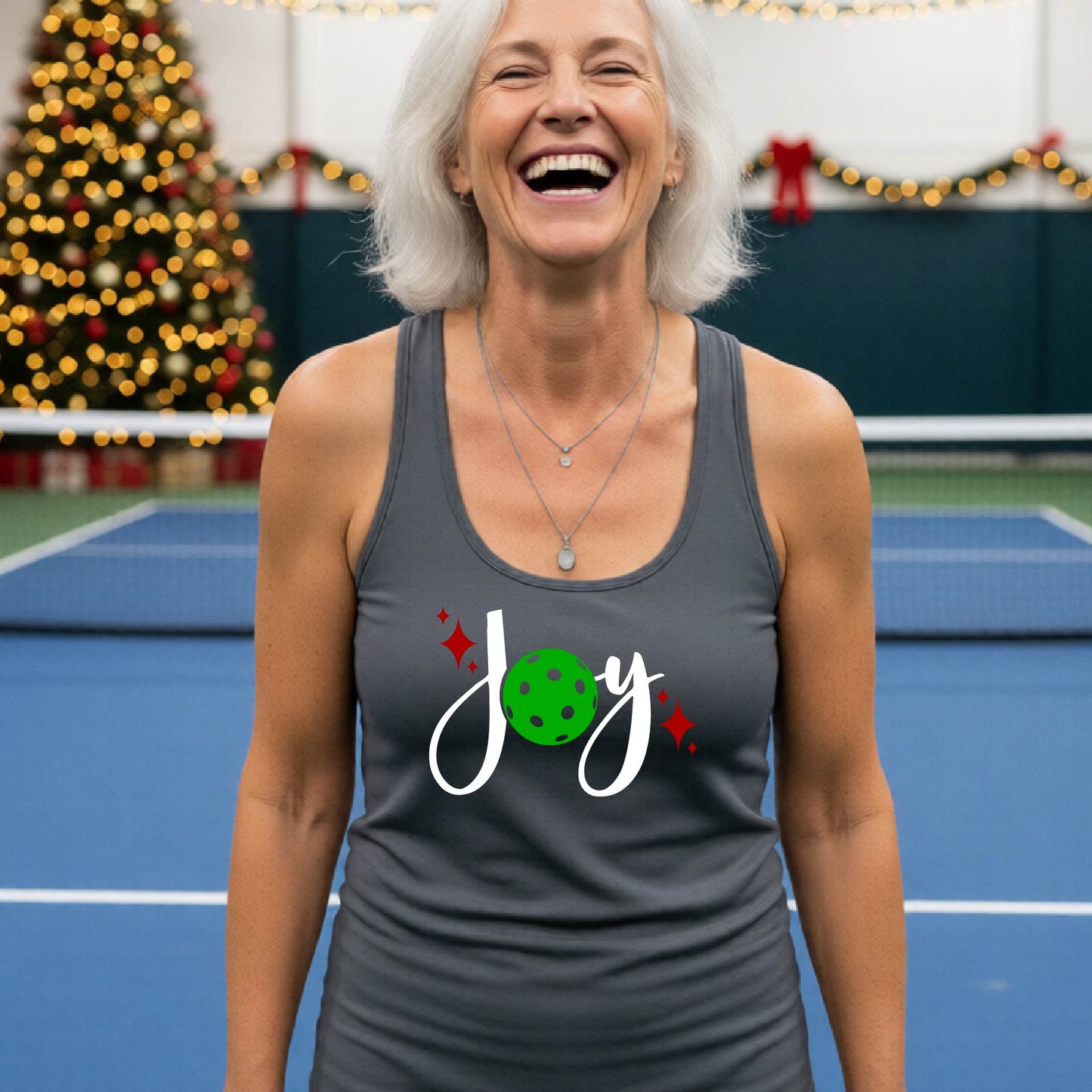 A woman laughing on an indoor pickleball court wearing a dark gray racerback tank top with a festive graphic that reads 'Joy' in white script, featuring a green Christmas picleball and red sparkles.