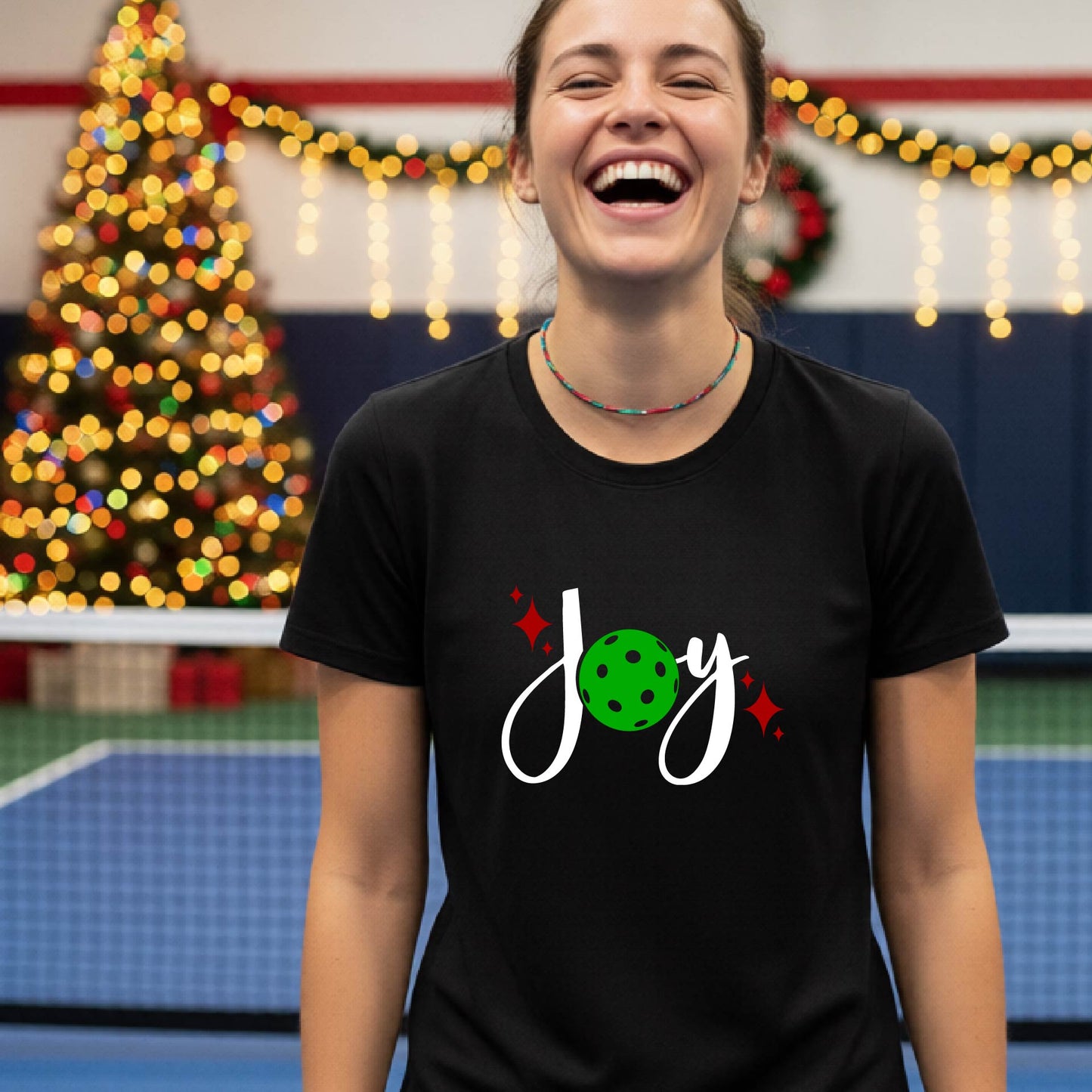 A woman wearing a black t-shirt with a 'Joy' graphic print featuring a green pickleball and white script, standing on an indoor pickleball court with Christmas decorations in the background.