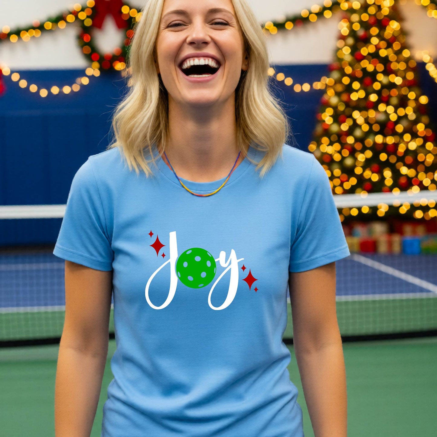 A woman wearing a light blue t-shirt with a 'Joy' graphic print featuring a green pickleball and white script, standing on an indoor pickleball court with Christmas decorations in the background.