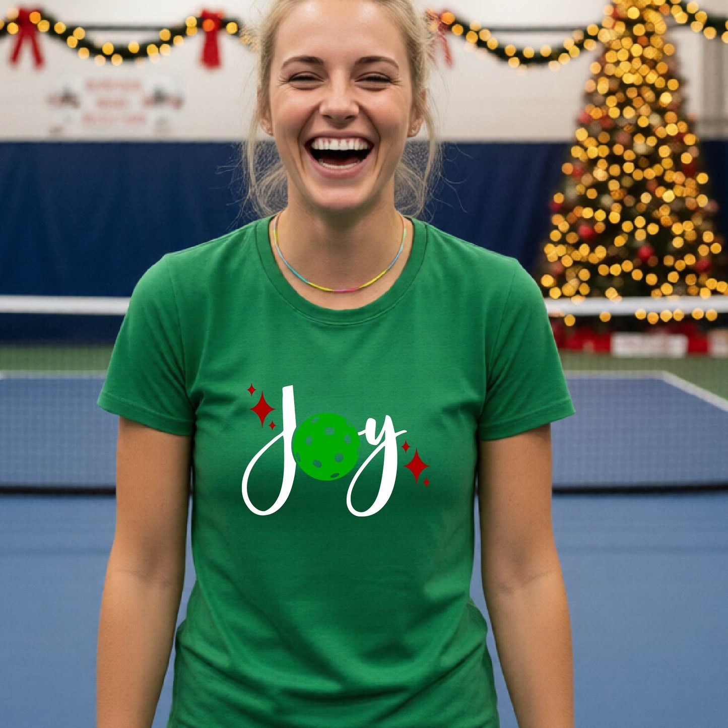 A woman wearing a green t-shirt with a 'Joy' graphic print featuring a green pickleball and white script, standing on an indoor pickleball court with Christmas decorations in the background.