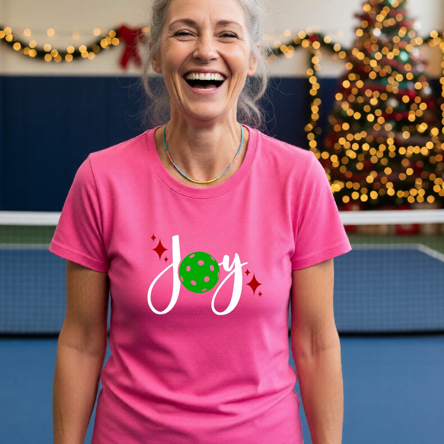 A woman wearing a pink t-shirt with a 'Joy' graphic print featuring a green pickleball and white script, standing on an indoor pickleball court with Christmas decorations in the background.