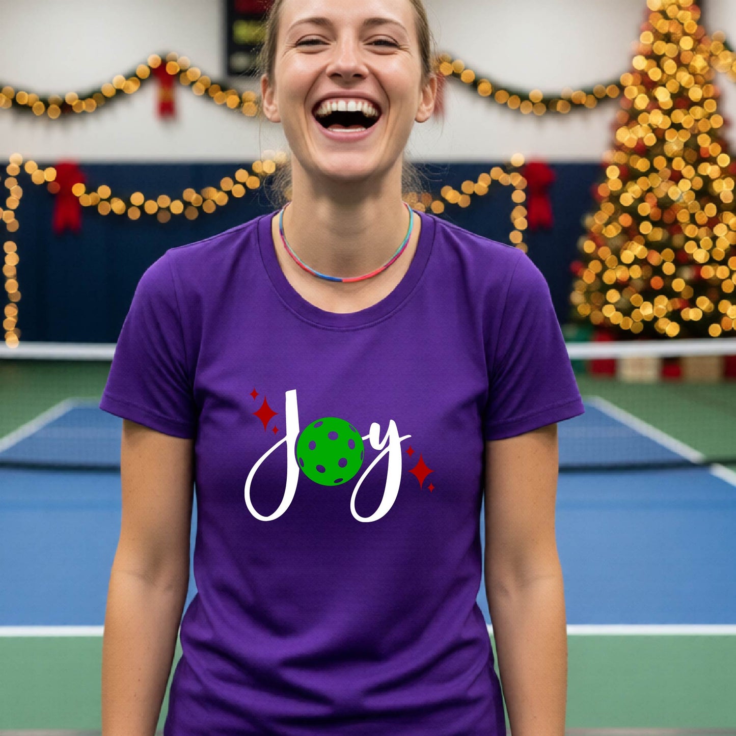 A woman wearing a purple t-shirt with a 'Joy' graphic print featuring a green pickleball and white script, standing on an indoor pickleball court with Christmas decorations in the background.