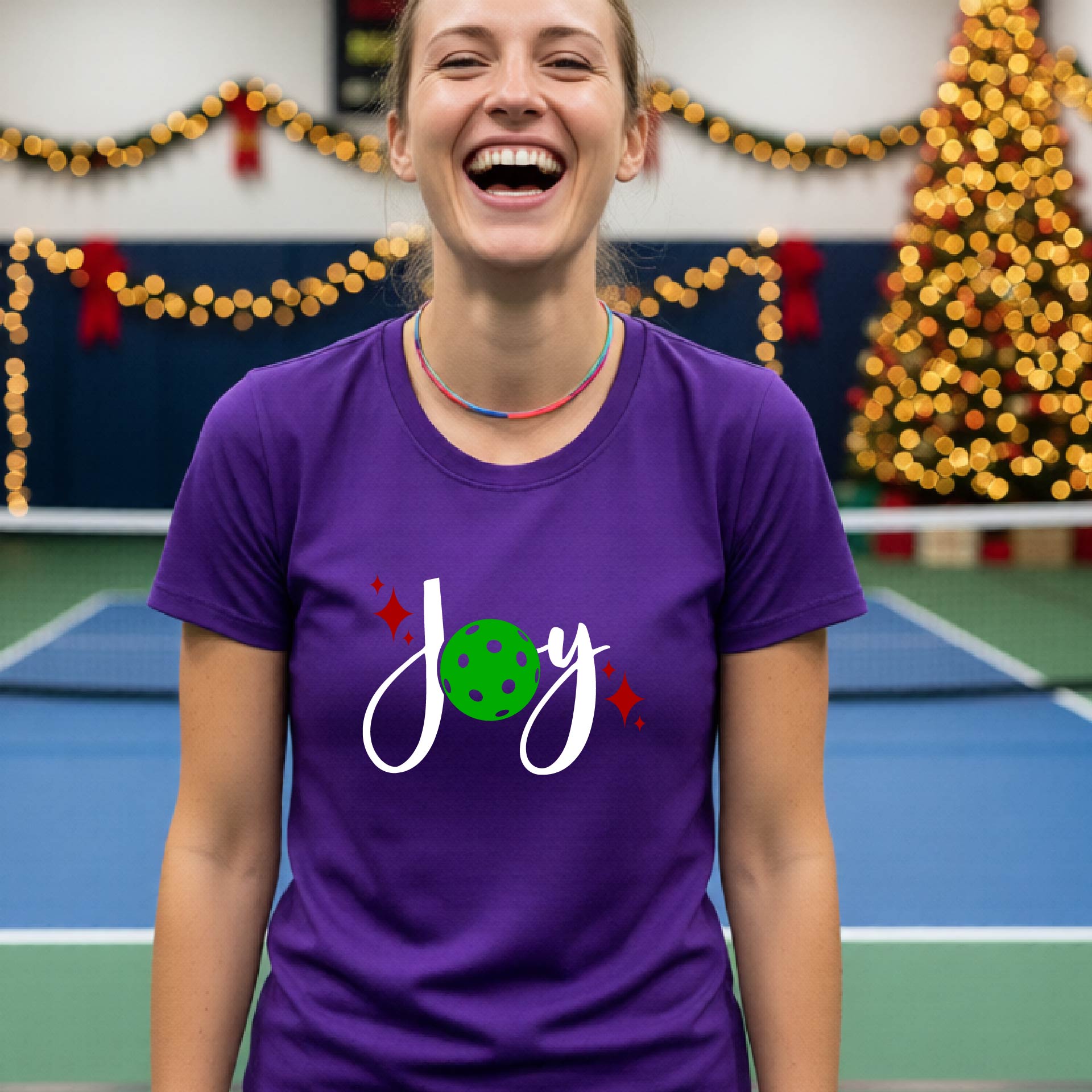 A woman wearing a purple t-shirt with a 'Joy' graphic print featuring a green pickleball and white script, standing on an indoor pickleball court with Christmas decorations in the background.