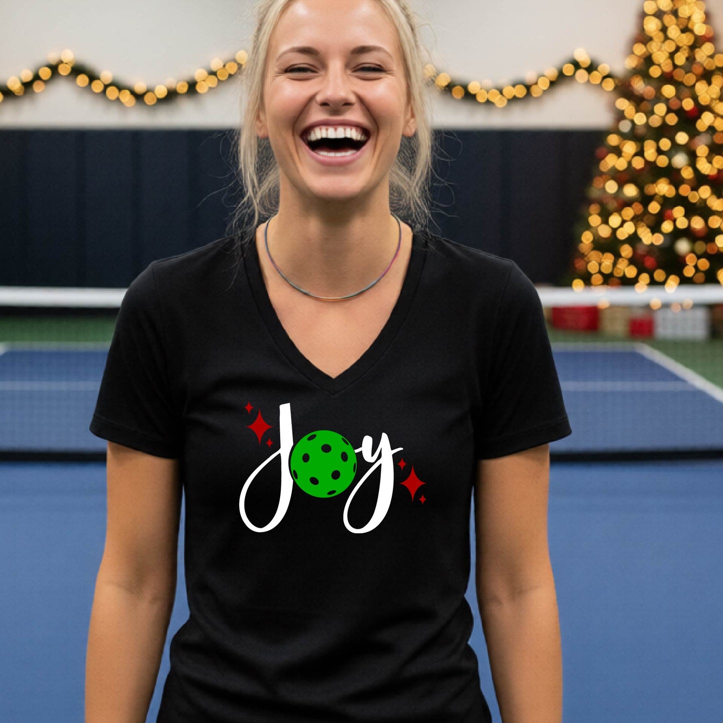 A woman smiling in a gym with a pickleball court  and a Christmas tree in the background, wearing a black v-neck t-shirt with a 'Joy' graphic featuring a green pickleball and white script.