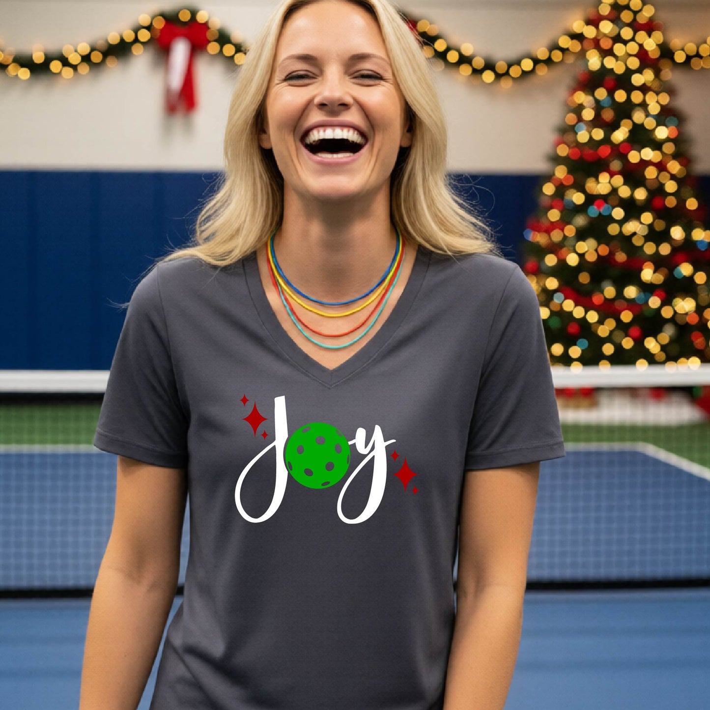 A woman smiling in a gym with a pickleball court  and a Christmas tree in the background, wearing a dark gray v-neck t-shirt with a 'Joy' graphic featuring a green pickleball and white script.